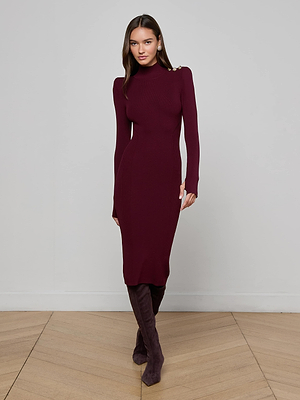 A woman stands against a light gray wall, wearing the L'AGENCE Larelle Midi Dress—a maroon ribbed knit with long sleeves, a mock neck, and shoulder buttons—paired with matching knee-high boots on a wooden floor.