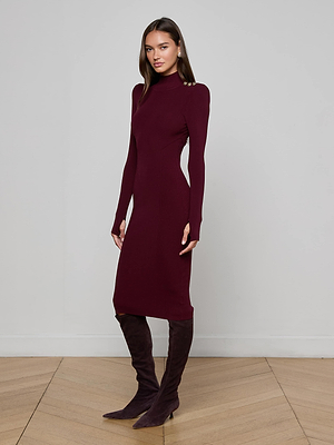 A woman poses on a light wood chevron floor against a plain gray wall, wearing the L'AGENCE Larelle Midi Dress—a ribbed knit, long-sleeve maroon dress—with matching knee-high boots, one foot slightly forward.