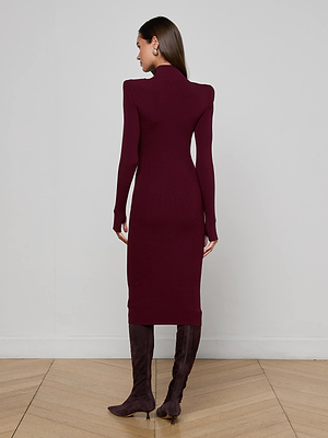 A woman faces away from the camera, wearing the L'AGENCE Larelle Midi Dress— a long-sleeved maroon ribbed knit—paired with knee-high dark brown boots, standing on a light wooden floor before a plain light gray wall.