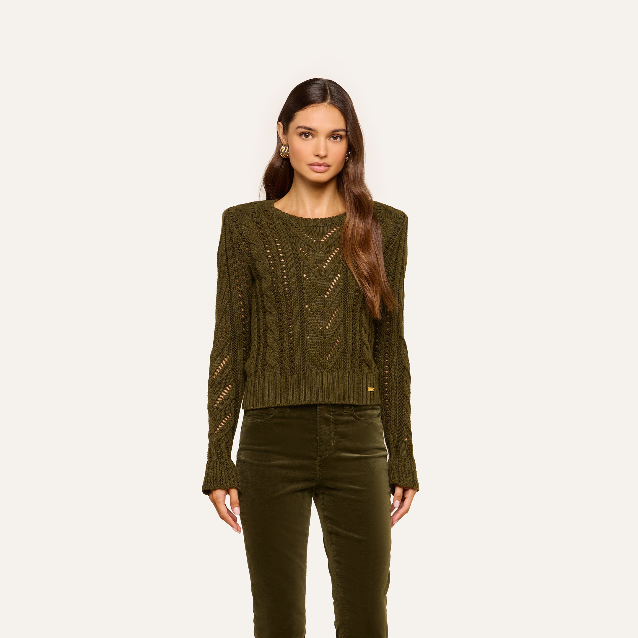 A woman with long brown hair wears the Mio Cabled Pullover in olive green and matching olive green pants, standing against a plain light background, facing forward with a neutral expression.