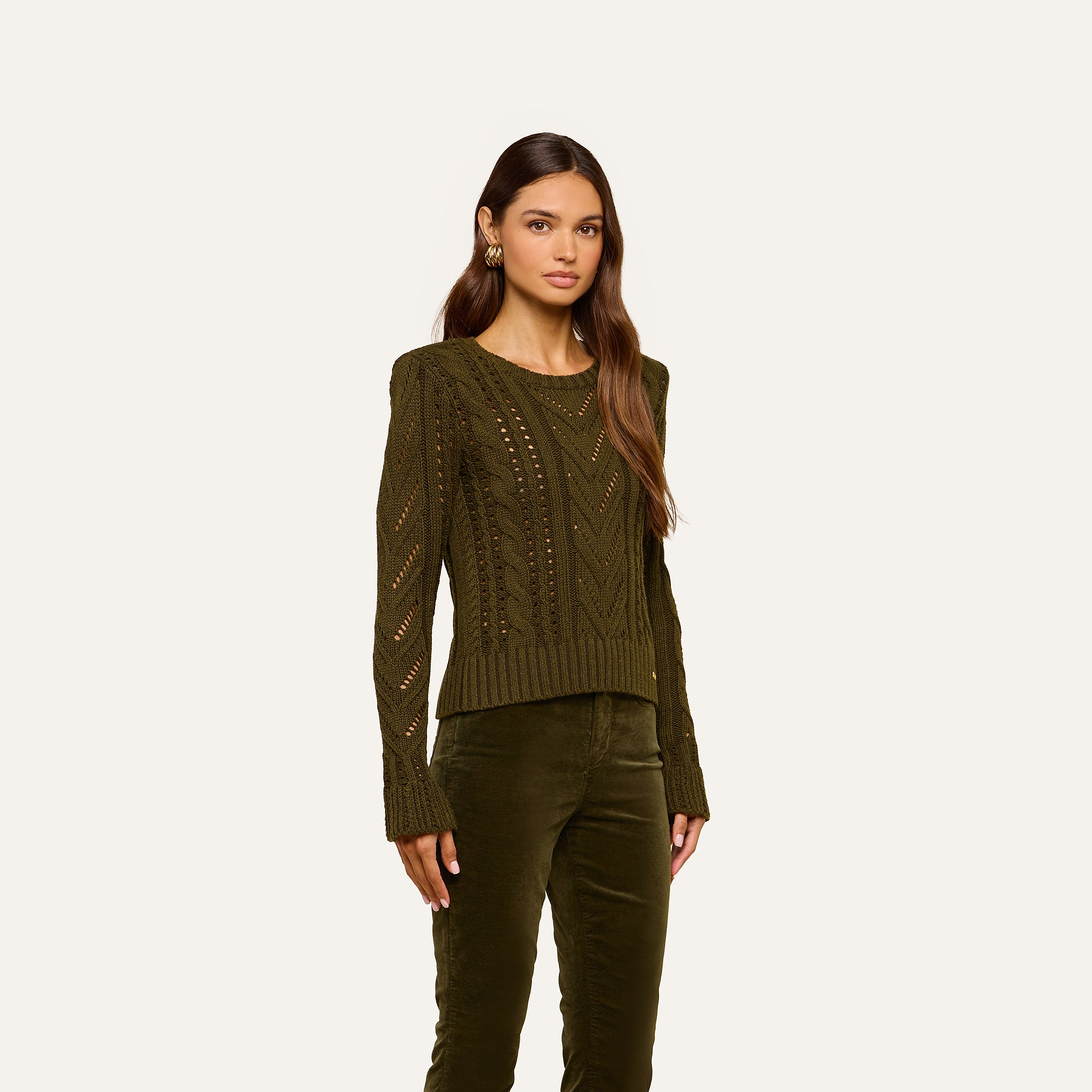 A woman with long, wavy hair and a neutral expression stands against a plain light background, wearing the Mio Cabled Pullover—an olive green cotton-blend knit sweater with long sleeves—paired with matching olive green pants.