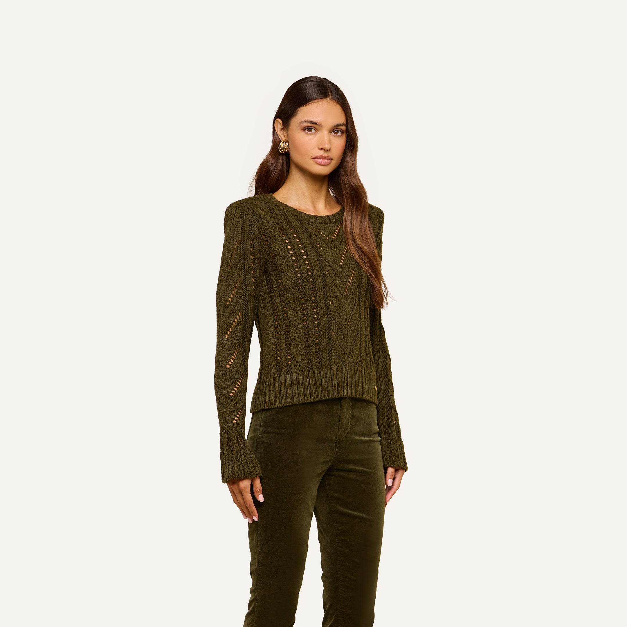 A woman with long, wavy hair and a neutral expression stands against a plain light background, wearing the Mio Cabled Pullover—an olive green cotton-blend knit sweater with long sleeves—paired with matching olive green pants.