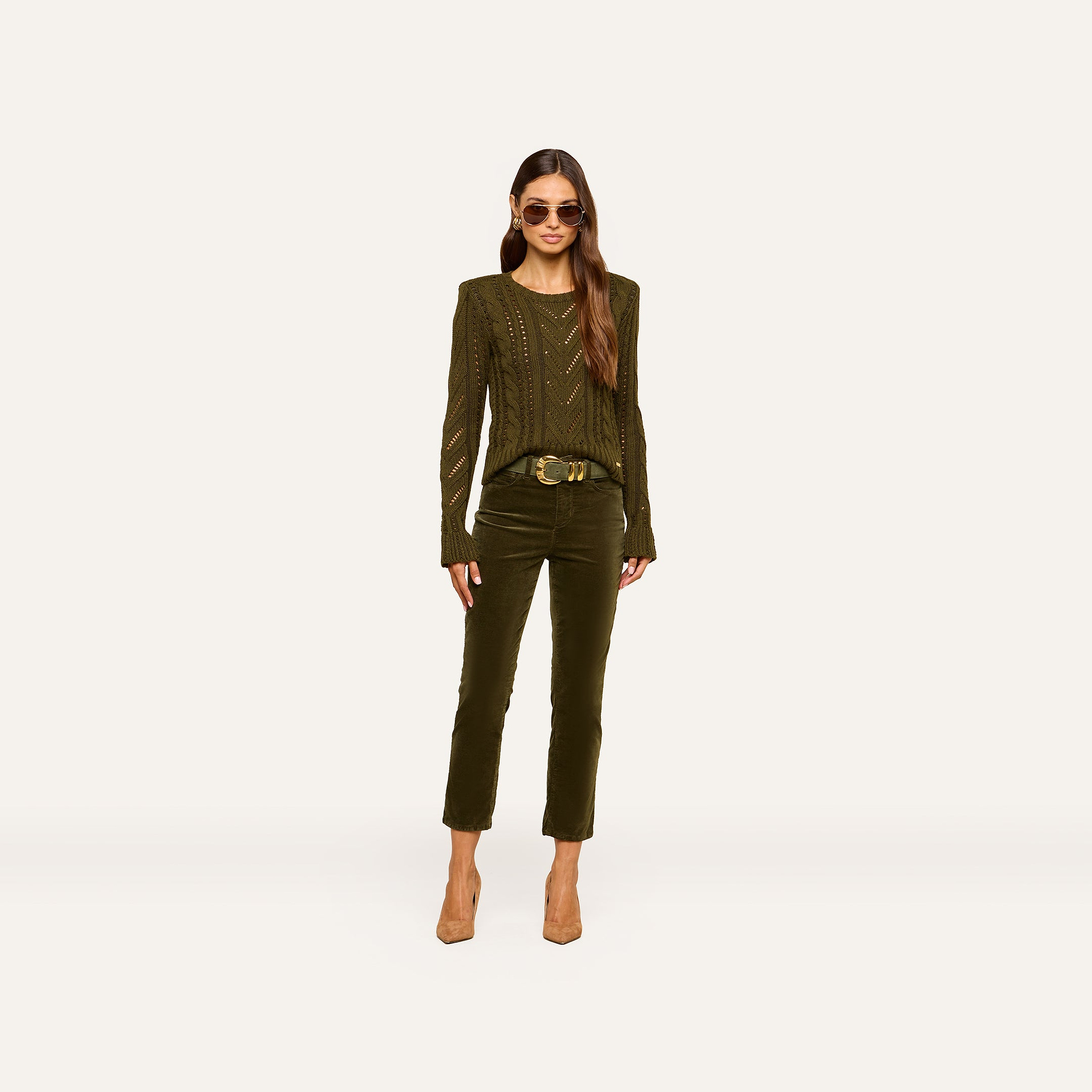 A woman wearing sunglasses, the Mio Cabled Pullover in olive green, matching pants, a belt, and beige heels stands against a plain white background.