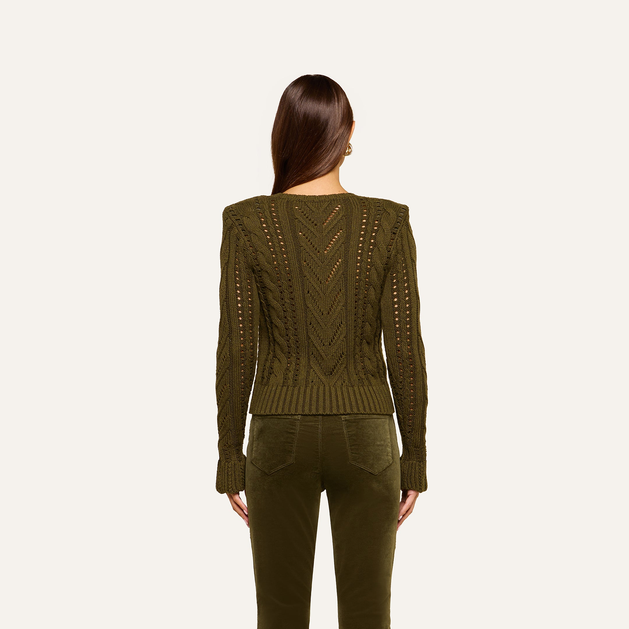 A woman with long brown hair, seen from behind, wears the Mio Cabled Pullover—a cotton-blend olive green crewneck sweater with a cabled knit—paired with matching green pants against a plain light background.