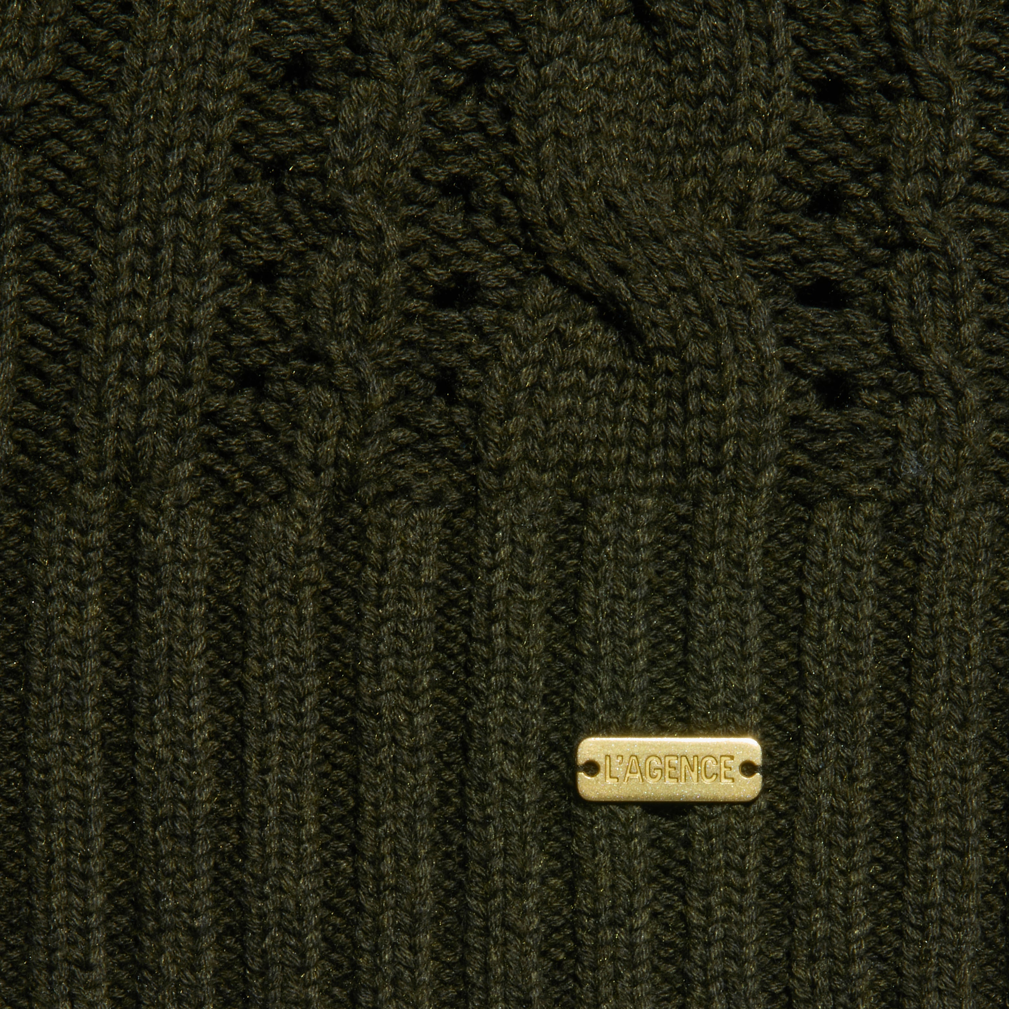 Close-up of the Mio Cabled Pullover, a dark green crewneck sweater crafted from a cotton-blend knit with ribbed and cable patterns. A gold metal tag engraved with LAGENCE accents the design.