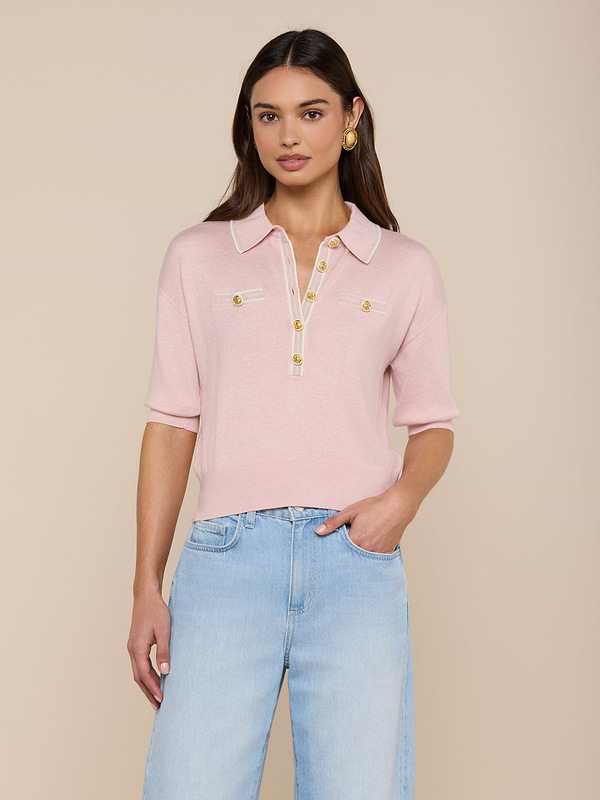 A woman stands against a plain white background wearing the Masaki Cropped Sweater—a light pink, featherweight knit polo top featuring gold buttons and front pockets—paired with light blue jeans.