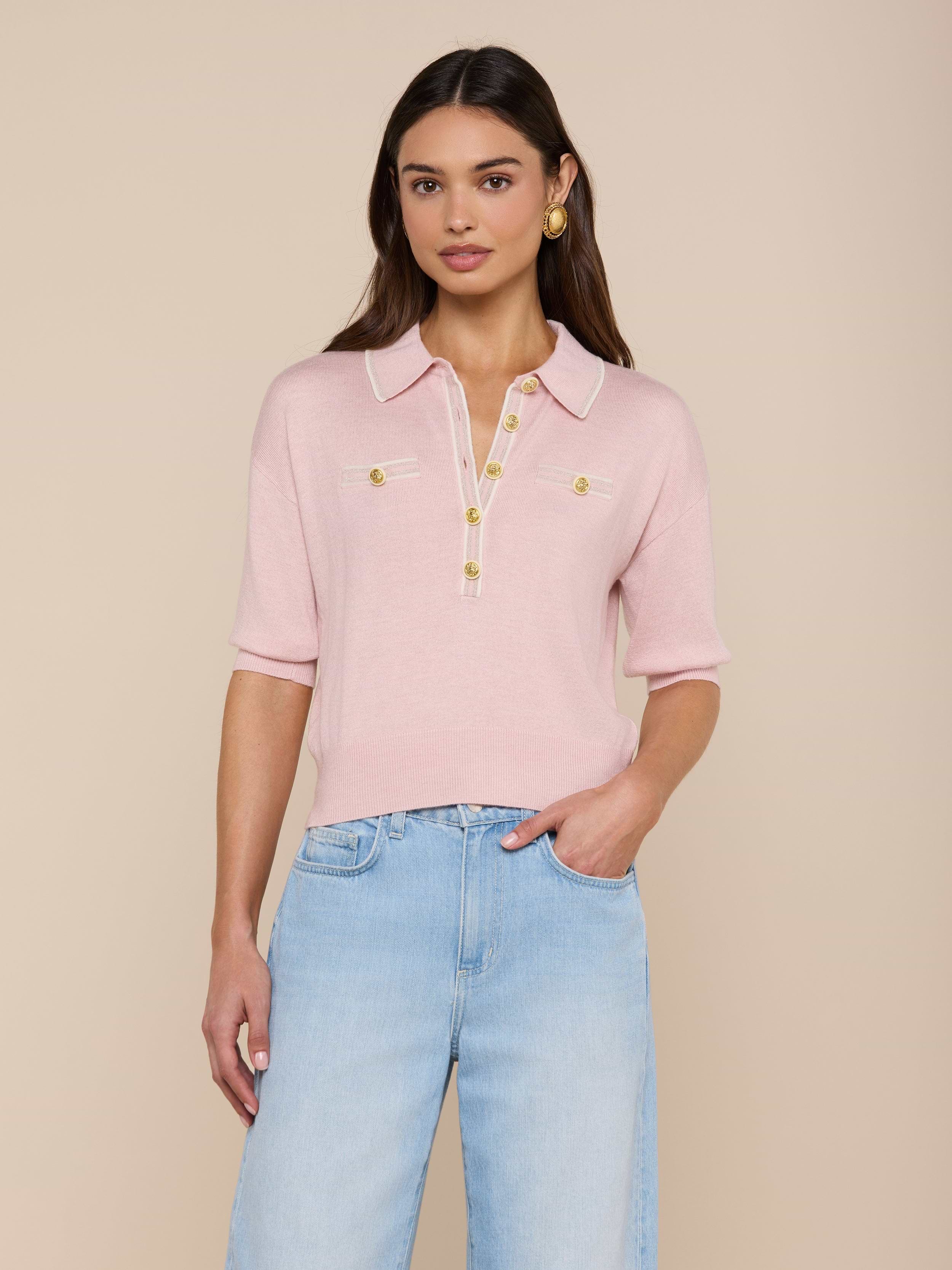 A woman stands against a plain white background wearing the Masaki Cropped Sweater—a light pink, featherweight knit polo top featuring gold buttons and front pockets—paired with light blue jeans.
