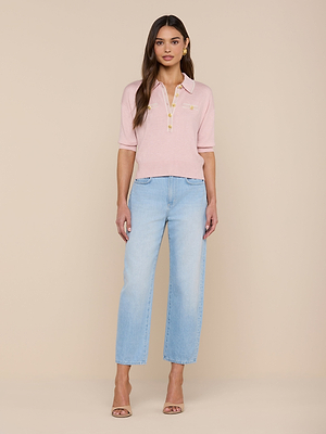 A woman stands against a beige background, wearing the Masaki Cropped Sweater in light pink with a zipper detail, paired with light blue high-waisted jeans and beige high-heeled sandals.