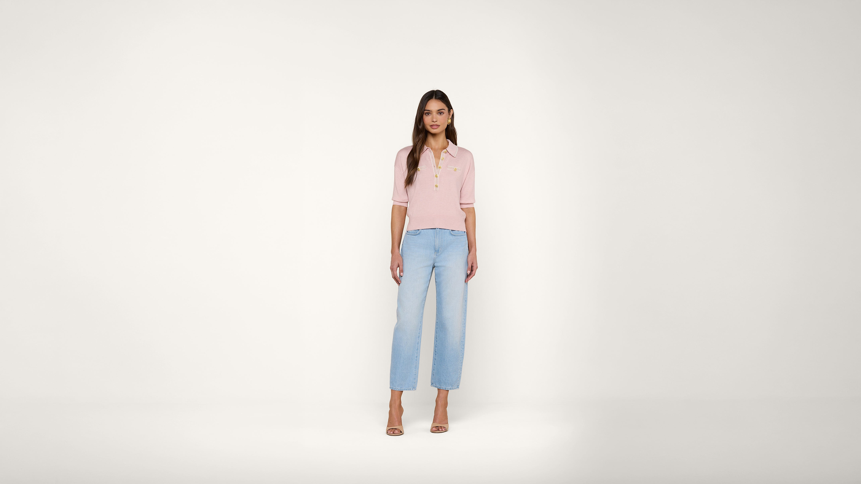 A woman with long brown hair stands facing forward, wearing a pink short-sleeve top, light blue wide-leg jeans, and beige heels against a plain white background.