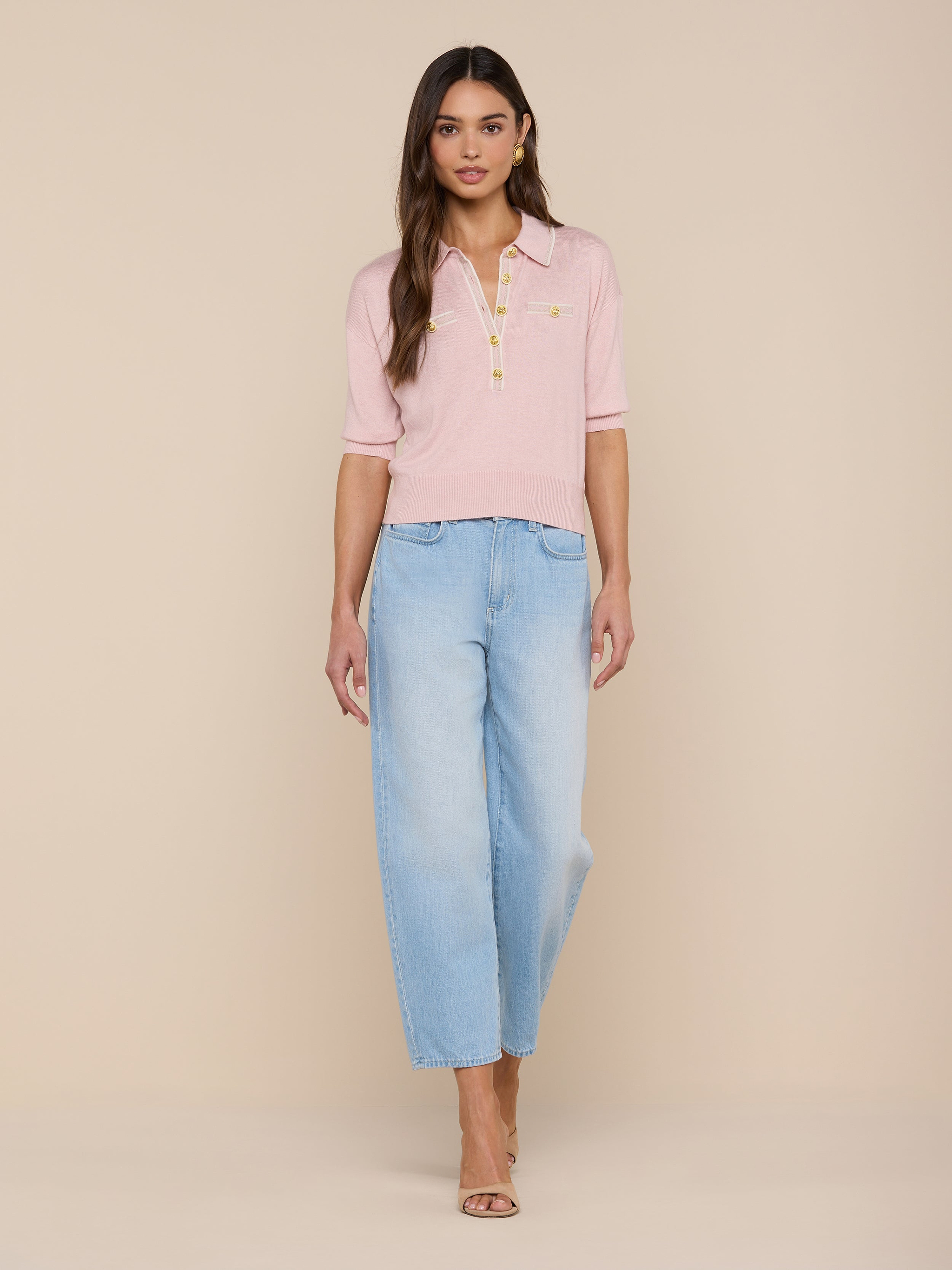 A woman stands facing forward, wearing the Masaki Cropped Sweater in light pink paired with light blue wide-leg jeans. She has long brown hair and is wearing open-toe heels against a plain white background.