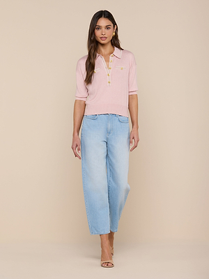 A woman with long brown hair and a neutral expression stands against a beige background, wearing the Masaki Cropped Sweater in light pink, paired with light blue wide-leg jeans and nude heeled sandals.