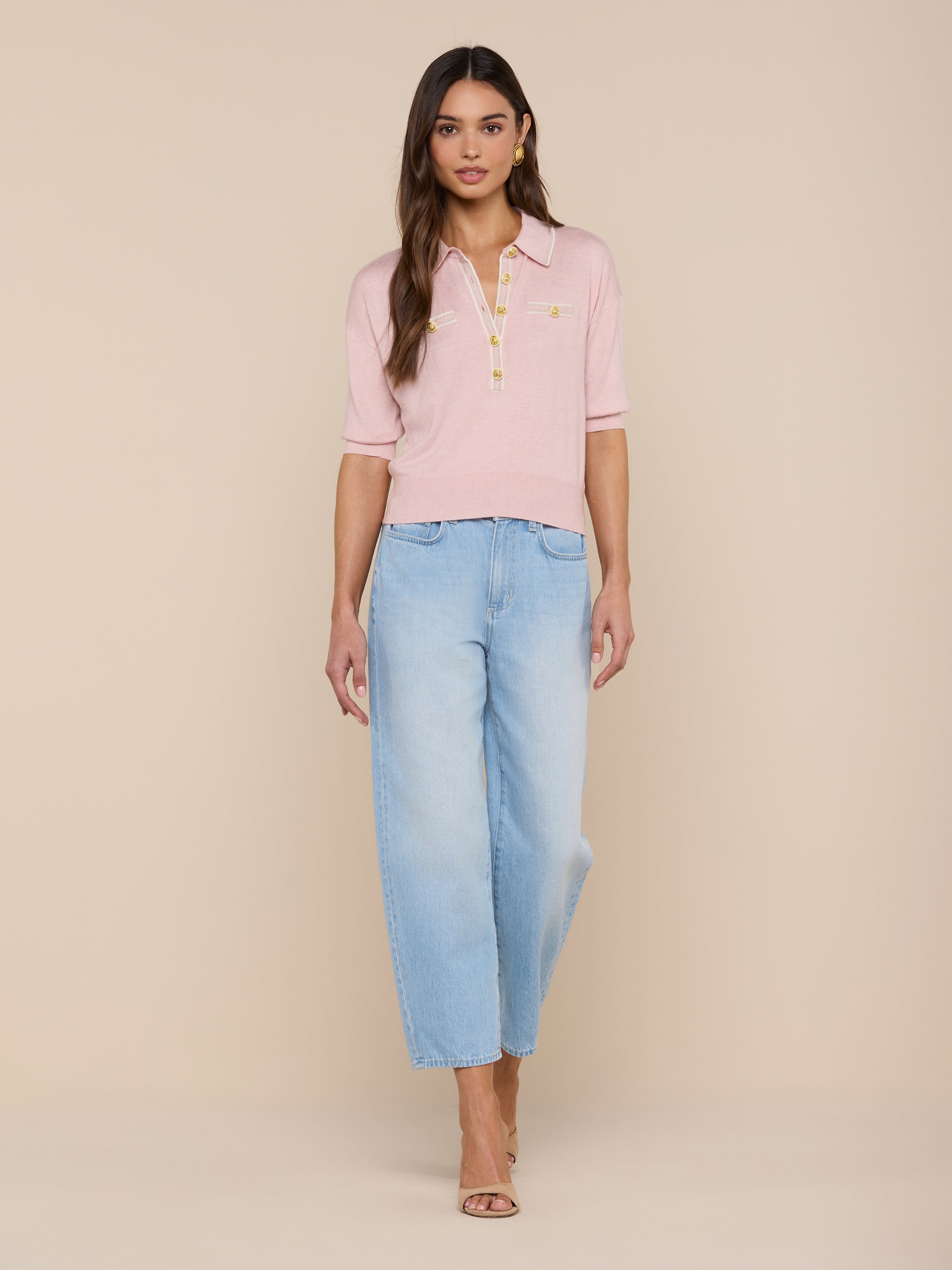 A woman stands facing forward, wearing the Masaki Cropped Sweater in light pink paired with light blue wide-leg jeans. She has long brown hair and is wearing open-toe heels against a plain white background.