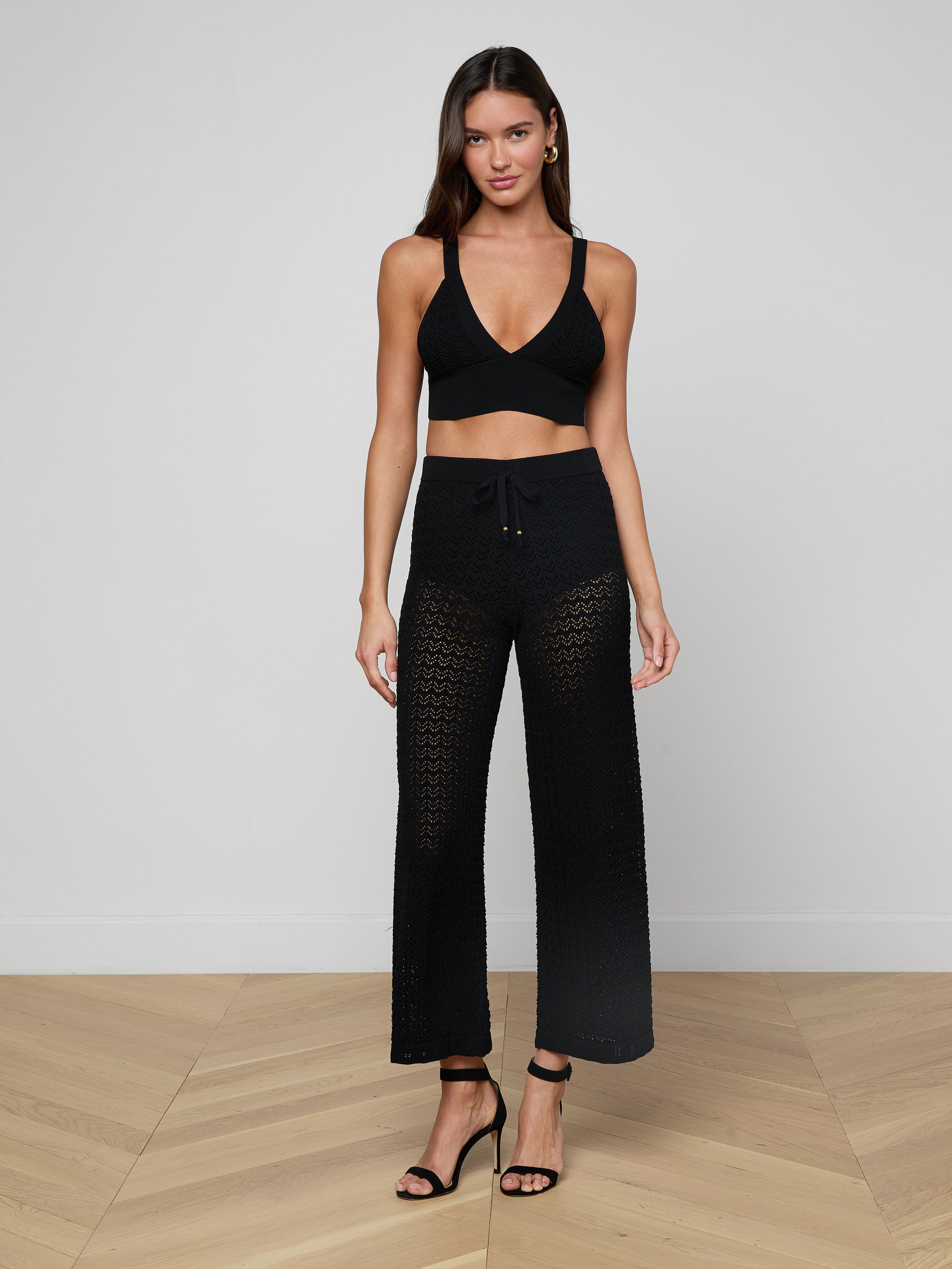 A woman stands against a plain background wearing the Reverie Pointelle Knit Bralette, which features a black hue and deep V-neckline. She pairs it with black crochet pants and black heeled sandals. Her long hair is down, and she poses in a relaxed stance with her arms at her sides.