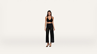 Woman standing against a plain white background, wearing a black halter crop top, black wide-leg mesh pants, and black high-heeled sandals. She has long brown hair and a neutral expression.