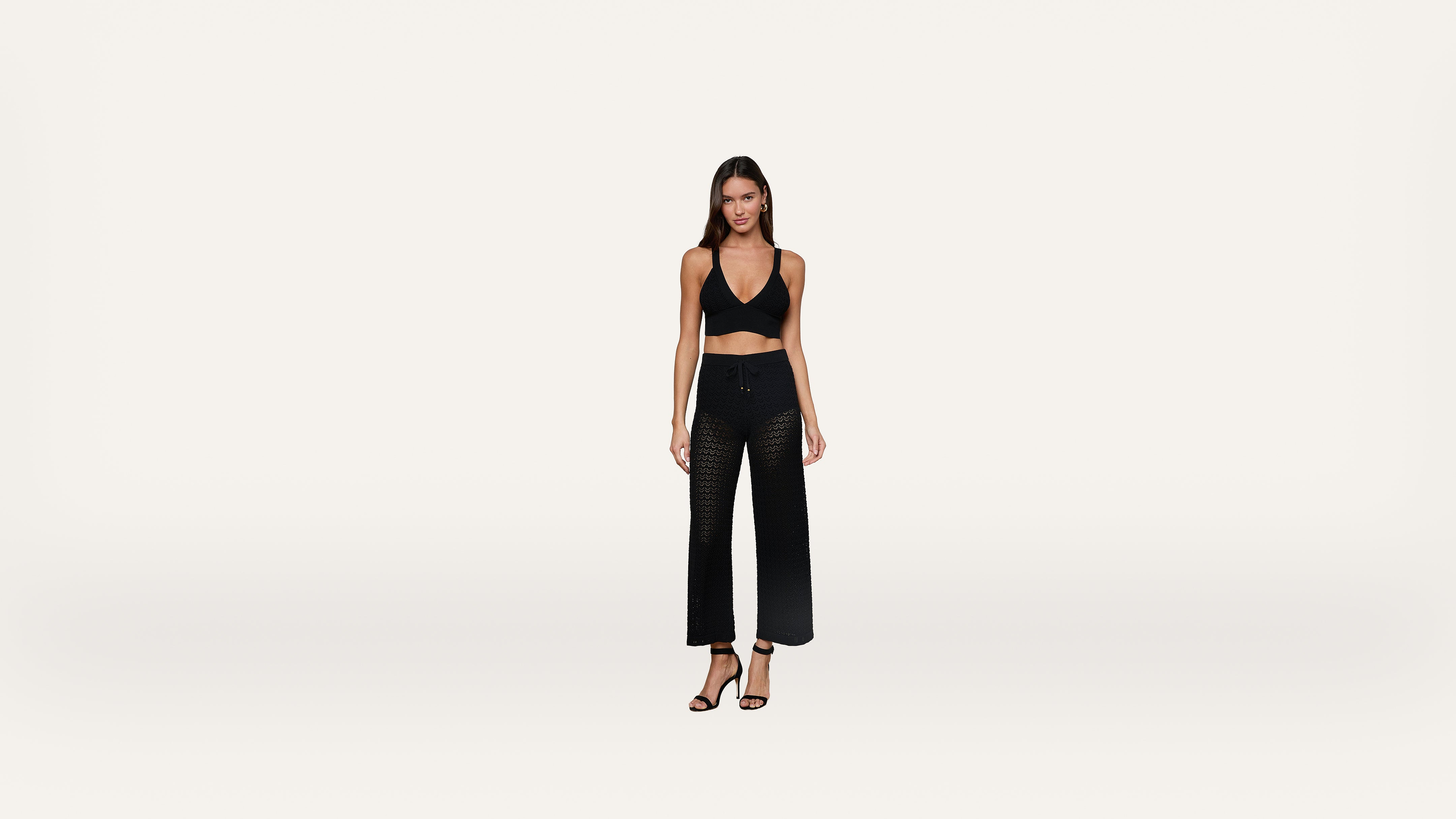Woman standing against a plain white background, wearing a black halter crop top, black wide-leg mesh pants, and black high-heeled sandals. She has long brown hair and a neutral expression.