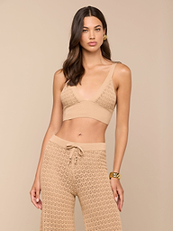 Una mujer con el pelo largo y castaño lleva el Reverie Pointelle Knit Bralette en color tostado con unos pantalones de cintura alta y pierna ancha a juego. Sobre un fondo beige, lleva pendientes dorados y una pulsera.