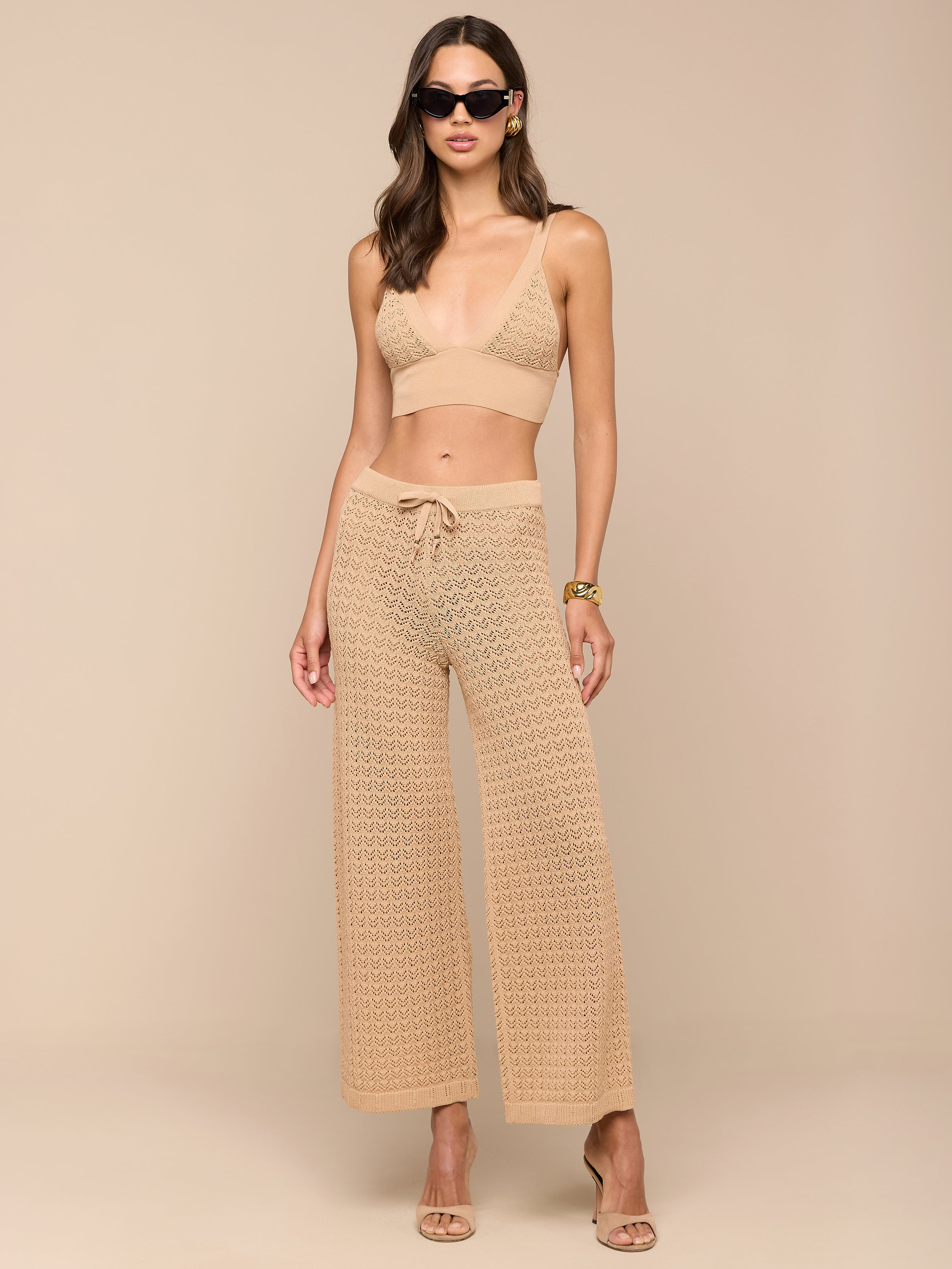 Una mujer con gafas de sol y el Reverie Pointelle Knit Bralette, combinado con unos pantalones anchos de crochet a juego, posa sobre un fondo beige. Completa su look con sandalias y una pulsera.