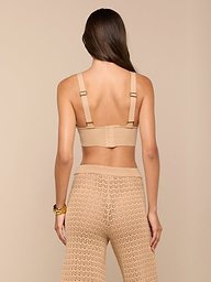 Una mujer de pelo largo y castaño aparece de espaldas con el Reverie Pointelle Knit Bralette en color beige y unos pantalones de cintura alta de punto de pointelle a juego, combinados con una gruesa pulsera dorada en la muñeca derecha.