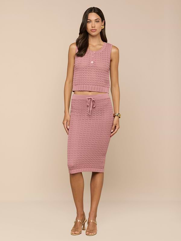 Una donna dai lunghi capelli scuri indossa la Lacen Pointelle Knit Skirt - una gonna midi in maglia rosa con coulisse - abbinata a un crop top rosa senza maniche. Su uno sfondo beige, la donna indossa tacchi beige e gioielli minimali.