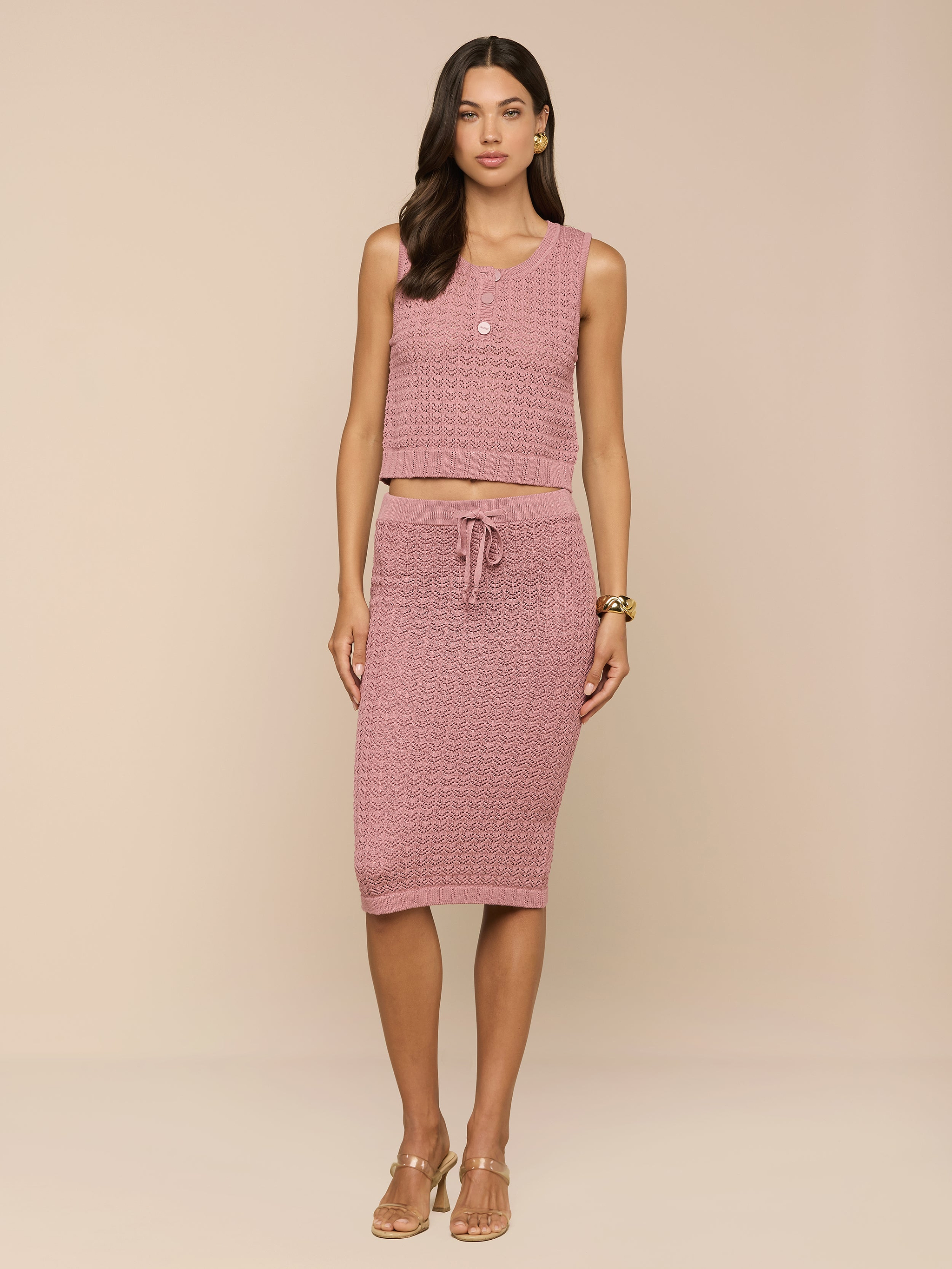 Una donna dai lunghi capelli scuri indossa la Lacen Pointelle Knit Skirt - una gonna midi in maglia rosa con coulisse - abbinata a un crop top rosa senza maniche. Su uno sfondo beige, la donna indossa tacchi beige e gioielli minimali.