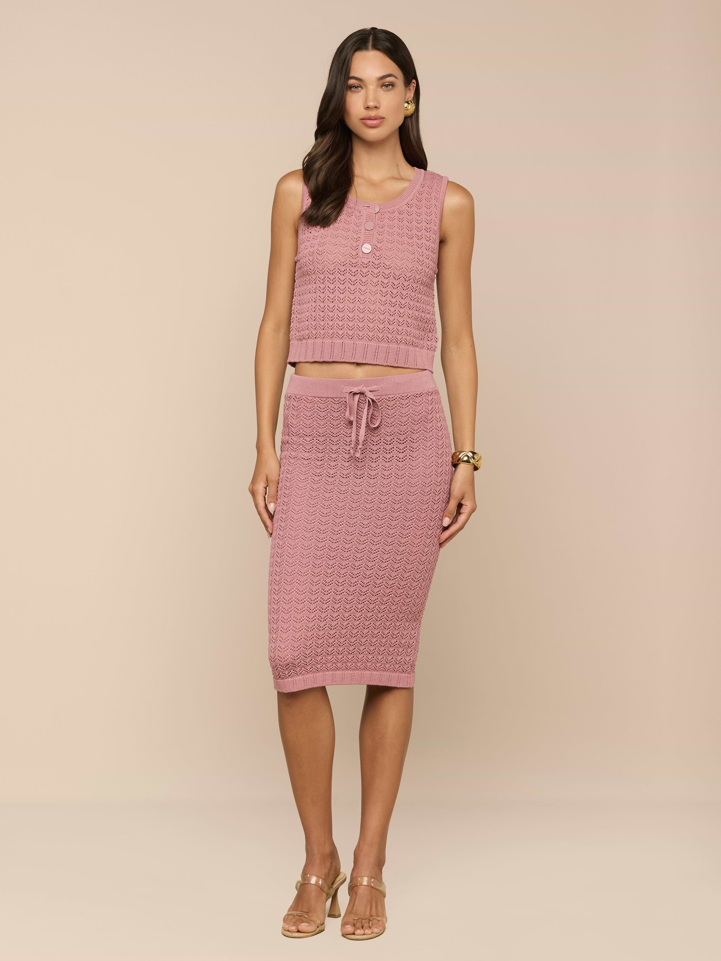 Una donna dai lunghi capelli scuri indossa la Lacen Pointelle Knit Skirt - una gonna midi in maglia rosa con coulisse - abbinata a un crop top rosa senza maniche. Su uno sfondo beige, la donna indossa tacchi beige e gioielli minimali.