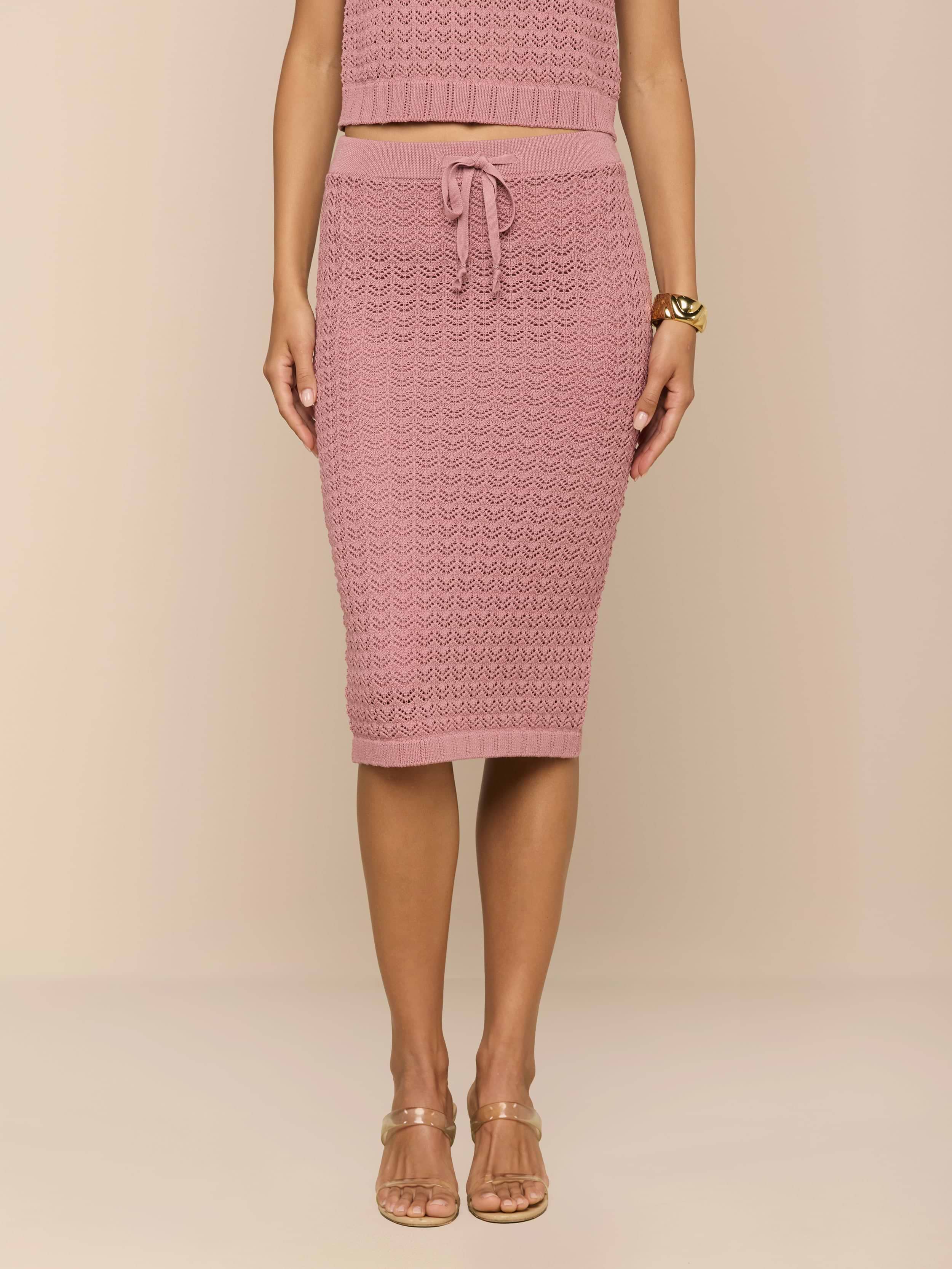 Una donna su uno sfondo beige indossa la Lacen Pointelle Knit Skirt in rosa, caratterizzata da una maglia traspirante, lunghezza midi, elastico in vita con coulisse, abbinata a un top cropped coordinato, bracciale dorato e tacchi con cinturino trasparente.