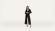A woman stands against a plain white background, wearing a black long-sleeve top and black wide-leg pants with high heels. She looks directly at the camera with her hands clasped.