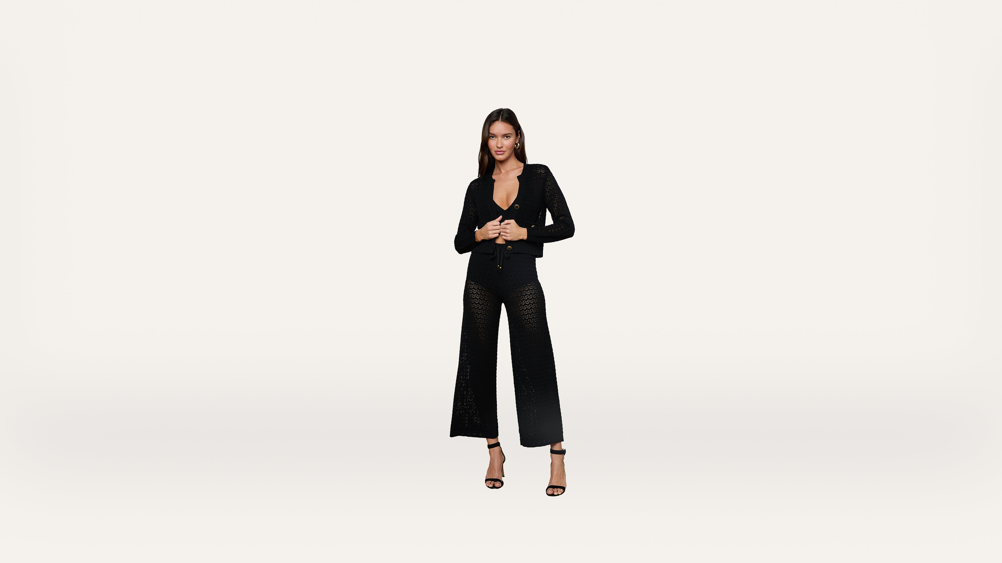 A woman stands against a plain white background, wearing a black long-sleeve top and black wide-leg pants with high heels. She looks directly at the camera with her hands clasped.