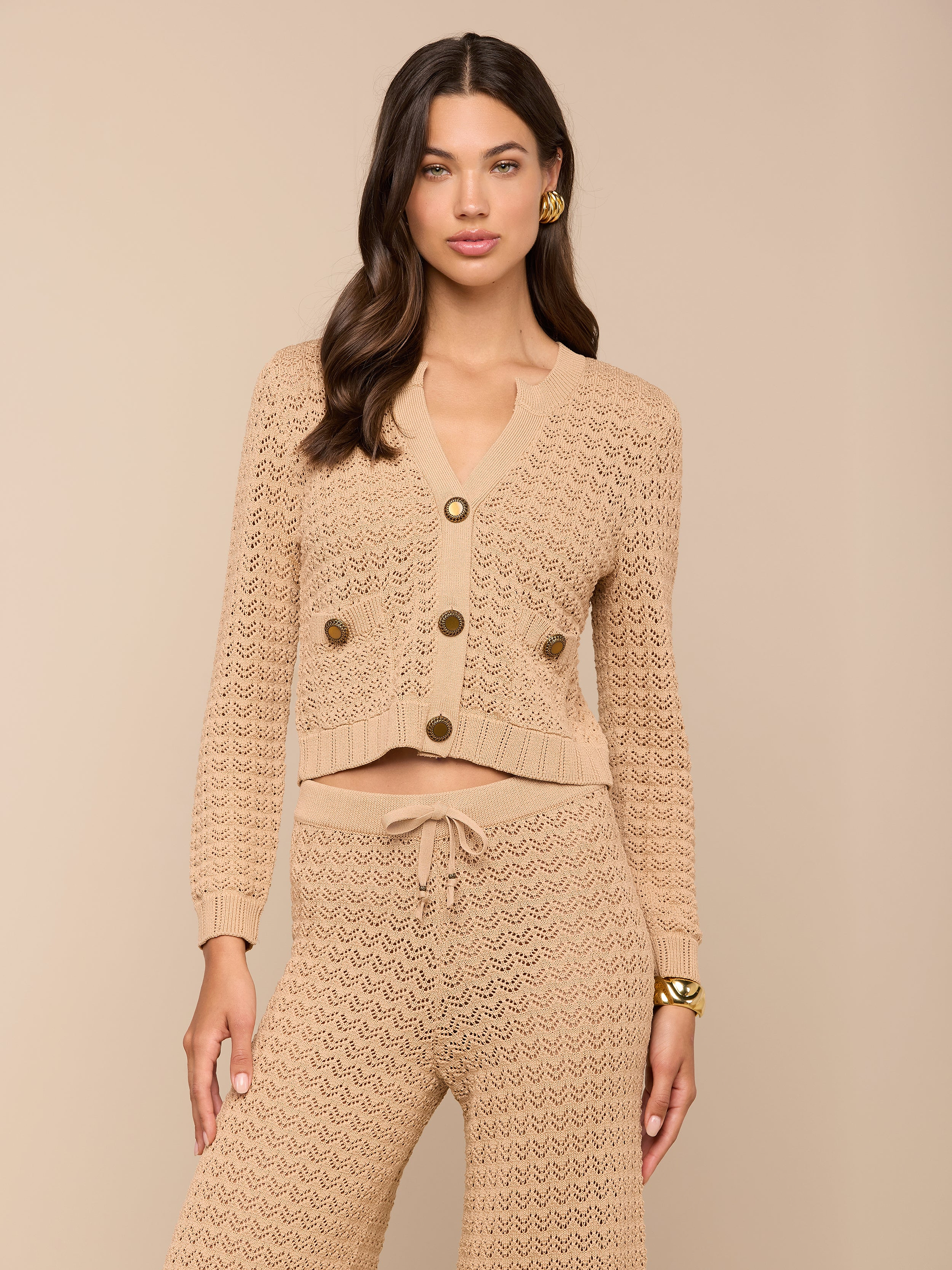 A woman models the Orla Pointelle Knit Cardigan in beige, featuring large buttons and paired with matching knit pants. She stands against a simple light background, her long wavy brown hair styled down and accessorized with gold earrings. Her neutral expression captures an effortless chic resort wear aesthetic.