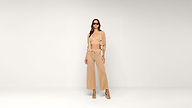 A woman in sunglasses wears a beige crochet outfit with a cropped top, cardigan, and wide-leg pants, standing against a plain white background.