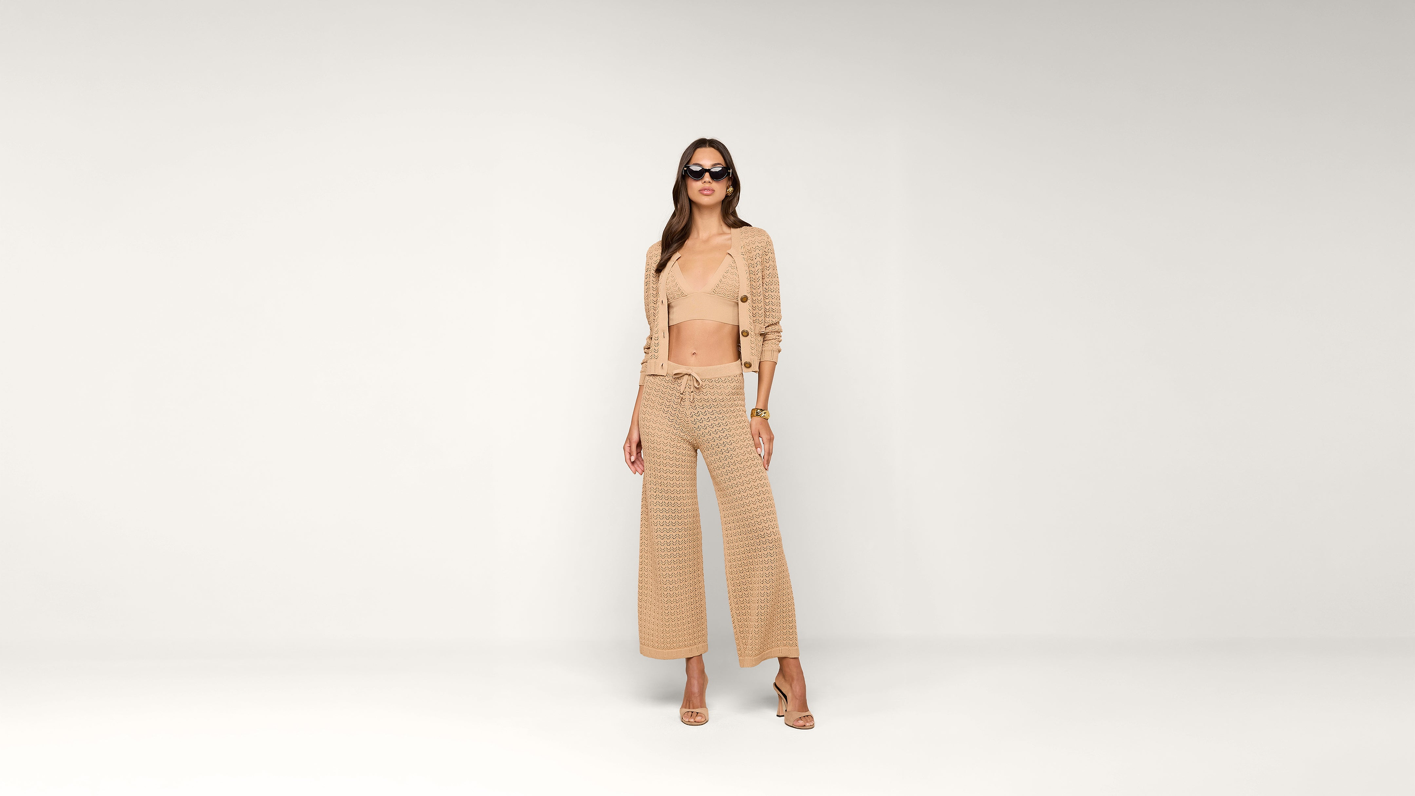 A woman in sunglasses wears a beige crochet outfit with a cropped top, cardigan, and wide-leg pants, standing against a plain white background.