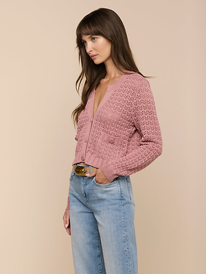A woman with long brown hair wears the Orla Pointelle Knit Cardigan in mauve and light blue jeans, standing against a beige background with one hand in her pocket, looking to the side.