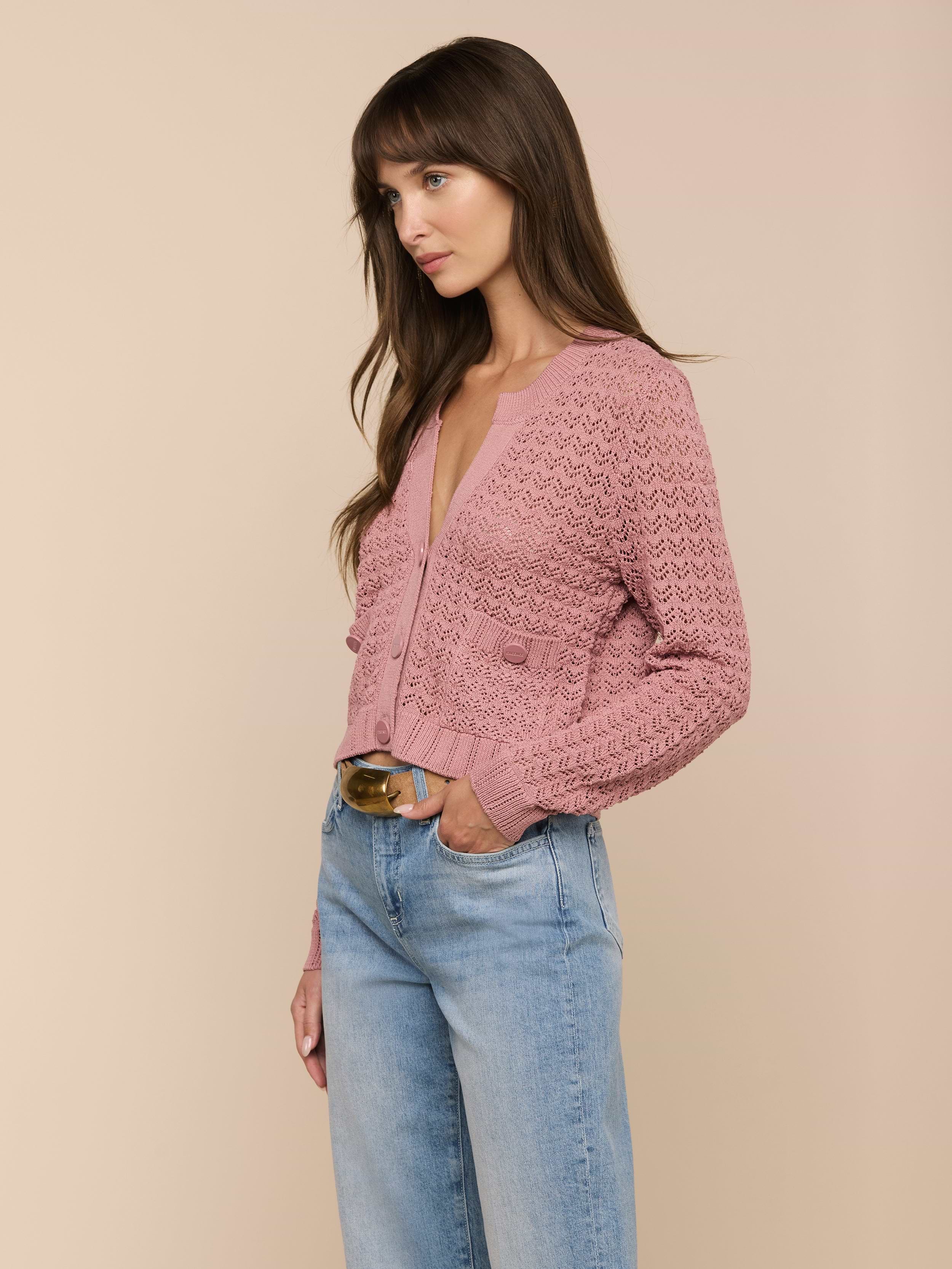Una donna con lunghi capelli castani indossa il cardigan Orla Pointelle Knit Cardigan color malva e jeans blu chiaro, in piedi su uno sfondo beige con una mano in tasca, guardando di lato.