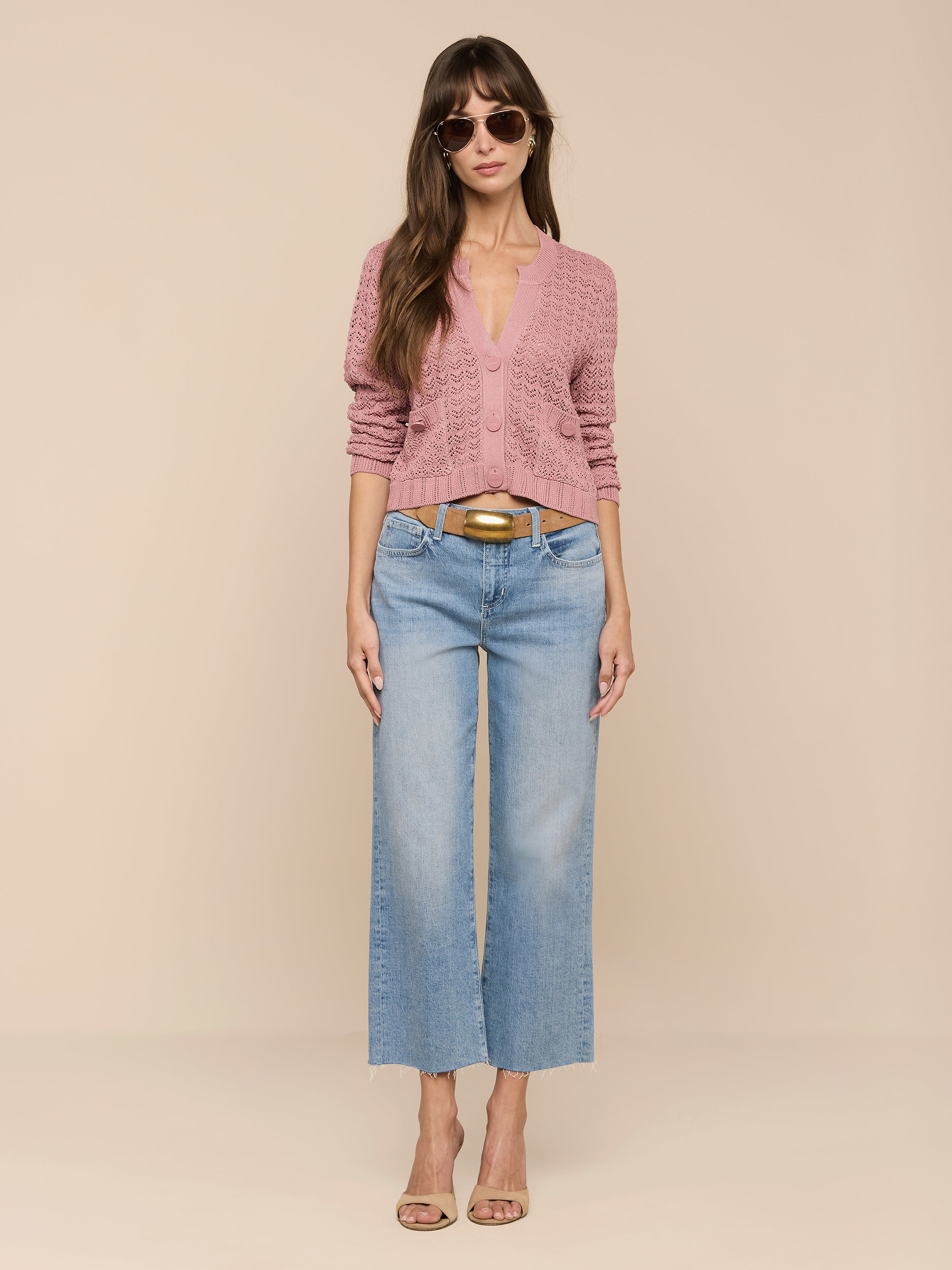 Una donna con lunghi capelli castani ondulati e una fibbia dorata indossa occhiali da sole, il cardigan Orla Pointelle Knit Cardigan color malva, jeans a gamba larga blu chiaro e tacchi beige su uno sfondo beige.