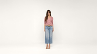 Woman wearing sunglasses, a pink long-sleeve top, blue high-waisted wide-leg jeans, and beige heels stands against a plain white background.