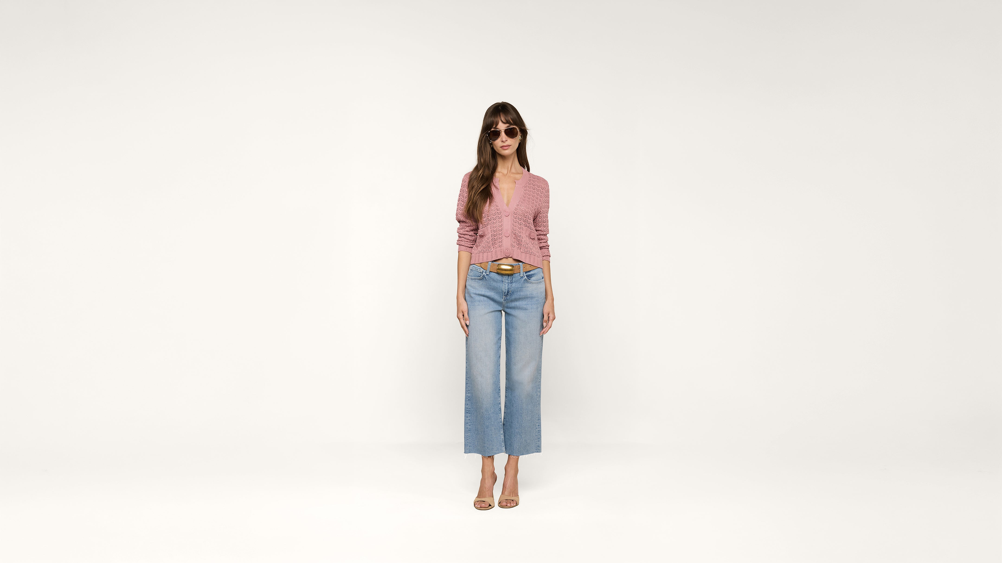 Woman wearing sunglasses, a pink long-sleeve top, blue high-waisted wide-leg jeans, and beige heels stands against a plain white background.
