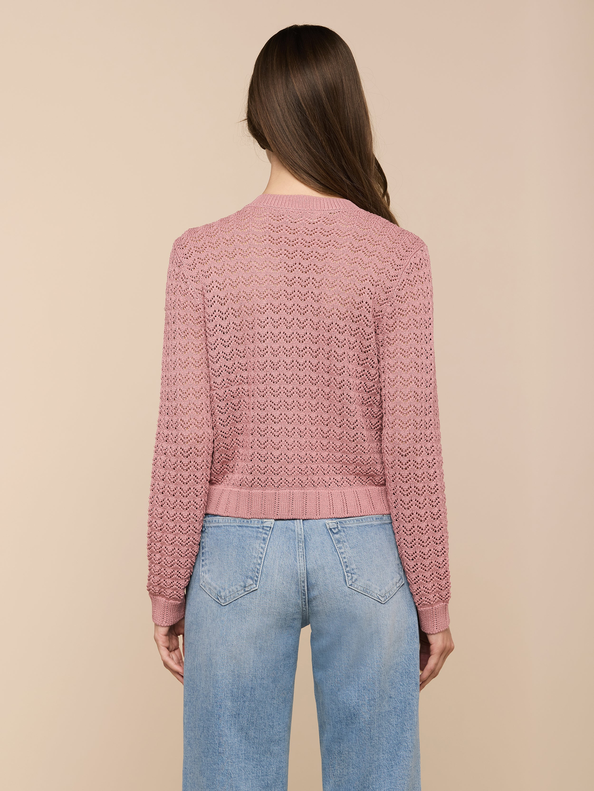 Una donna dai lunghi capelli castani è in piedi, rivolta verso lesterno, con indosso il cardigan Orla Pointelle Knit Cardigan color malva e jeans blu chiaro su uno sfondo beige.