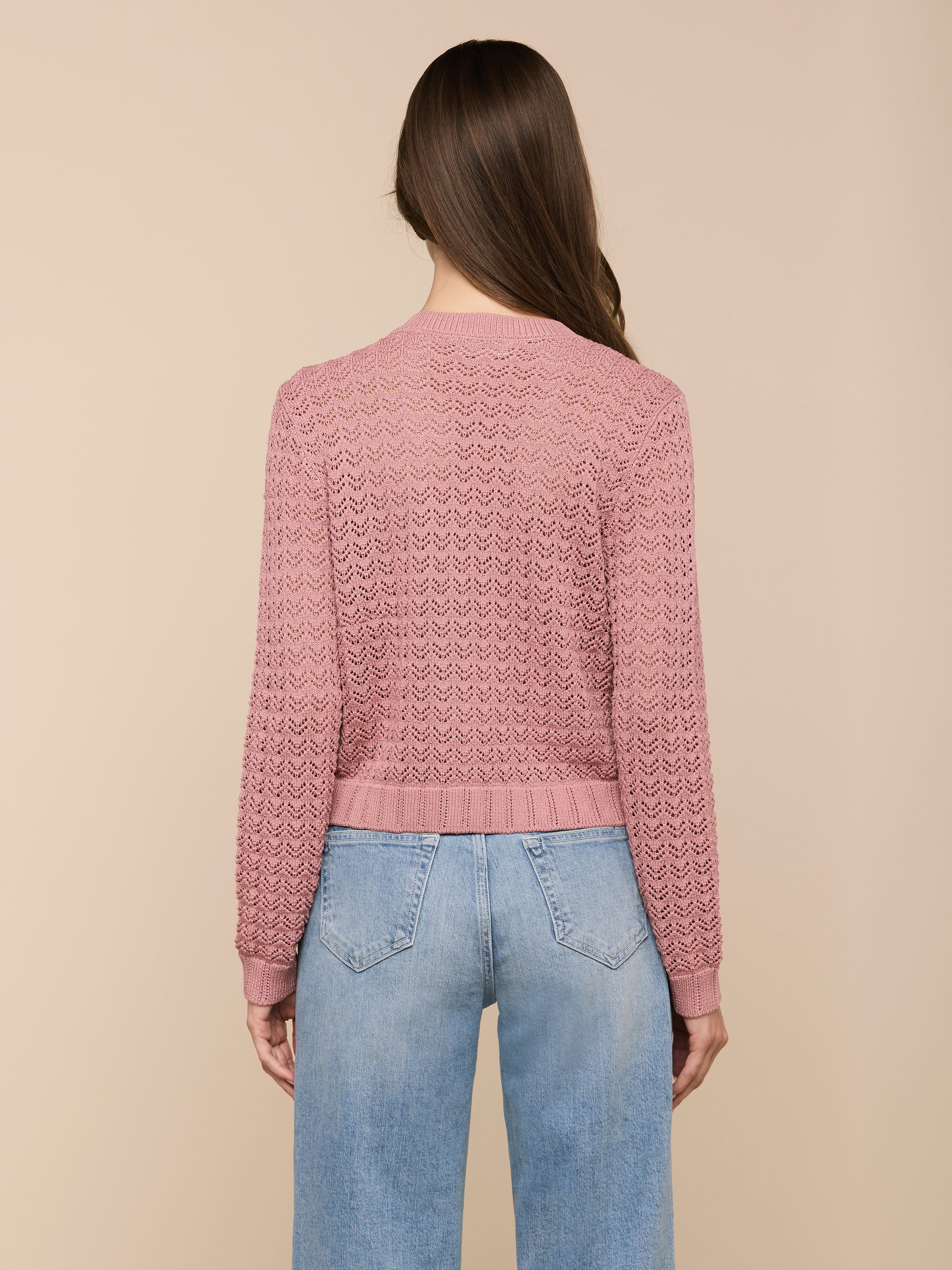 Una donna dai lunghi capelli castani è in piedi, rivolta verso lesterno, con indosso il cardigan Orla Pointelle Knit Cardigan color malva e jeans blu chiaro su uno sfondo beige.