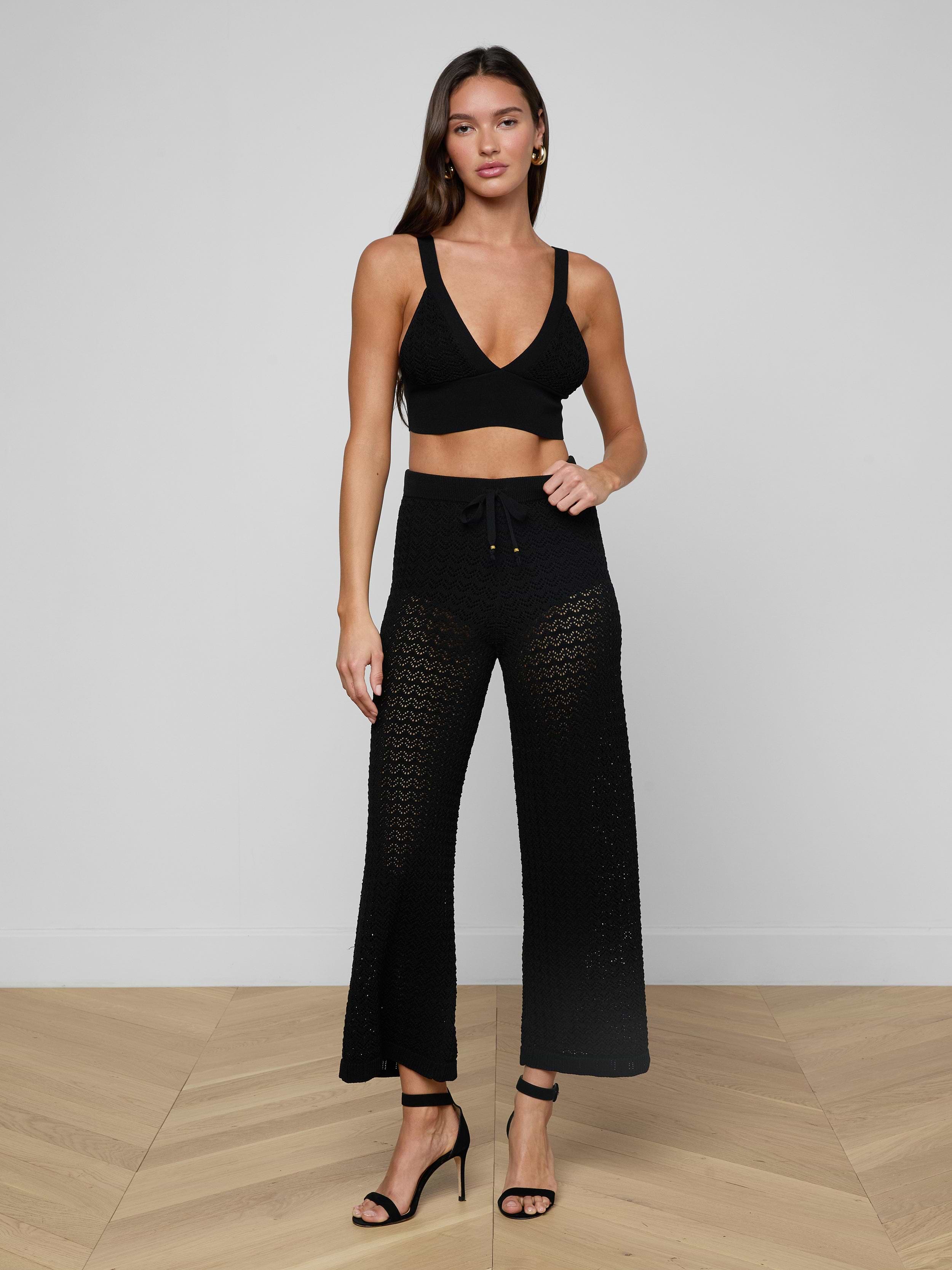A woman stands against a plain white background wearing a black crop top, the Gaelle Pointelle Knit Pant in black featuring a sheer crochet knit and drawstring waist, and black high-heeled sandals.