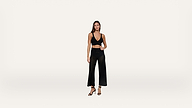 A woman stands against a plain white background, wearing a black sleeveless crop top, black high-waisted mesh wide-leg pants, and black heeled sandals.