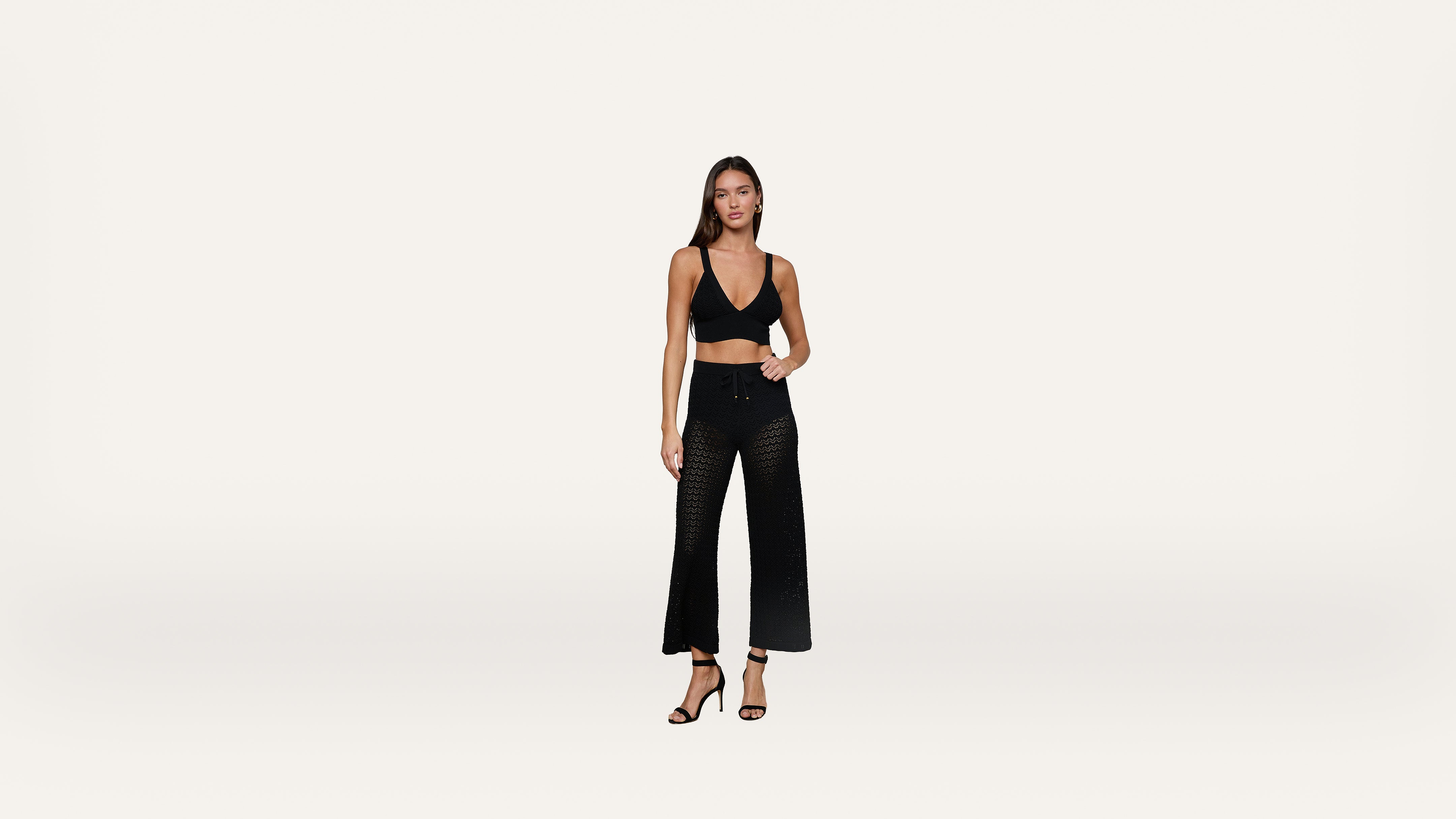 A woman stands against a plain white background, wearing a black sleeveless crop top, black high-waisted mesh wide-leg pants, and black heeled sandals.