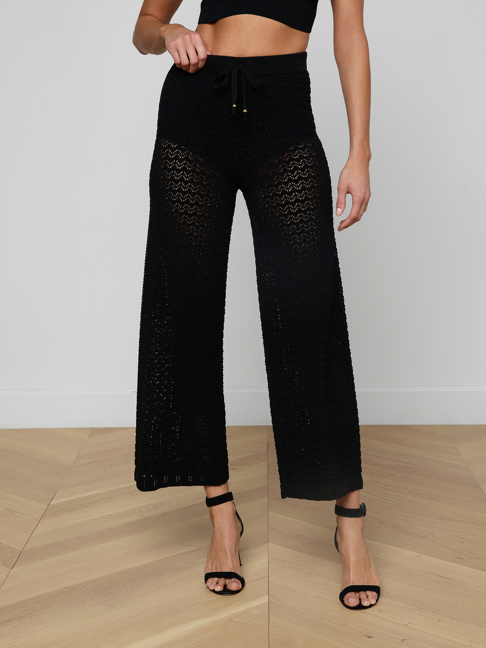 A woman wears the Gaelle Pointelle Knit Pant—a black, high-waisted, wide-leg knit pant with a drawstring waist—paired with a black crop top and black high-heeled sandals. Her face is not visible.