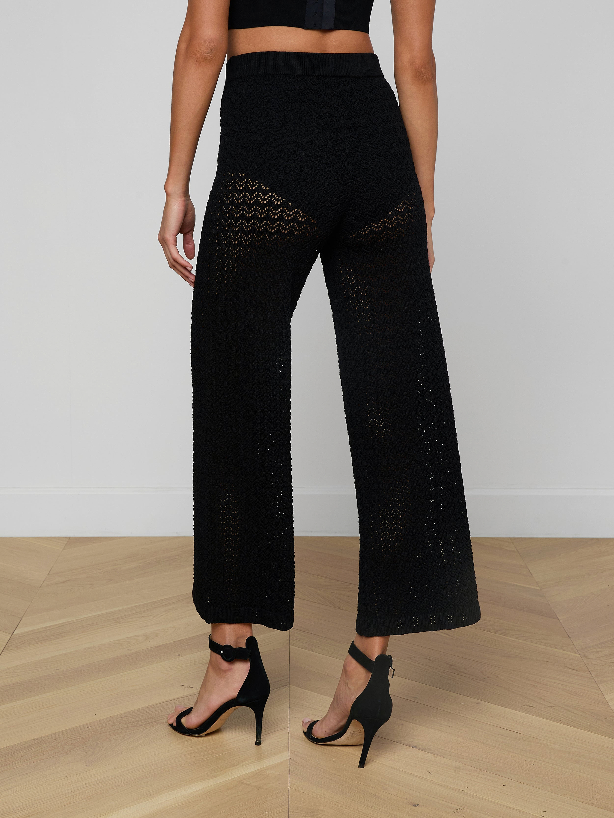 A person wearing Gaelle Pointelle Knit Pants in black, featuring a wide-leg silhouette and drawstring waist, paired with black high-heeled sandals and shown from the back against a plain light background.