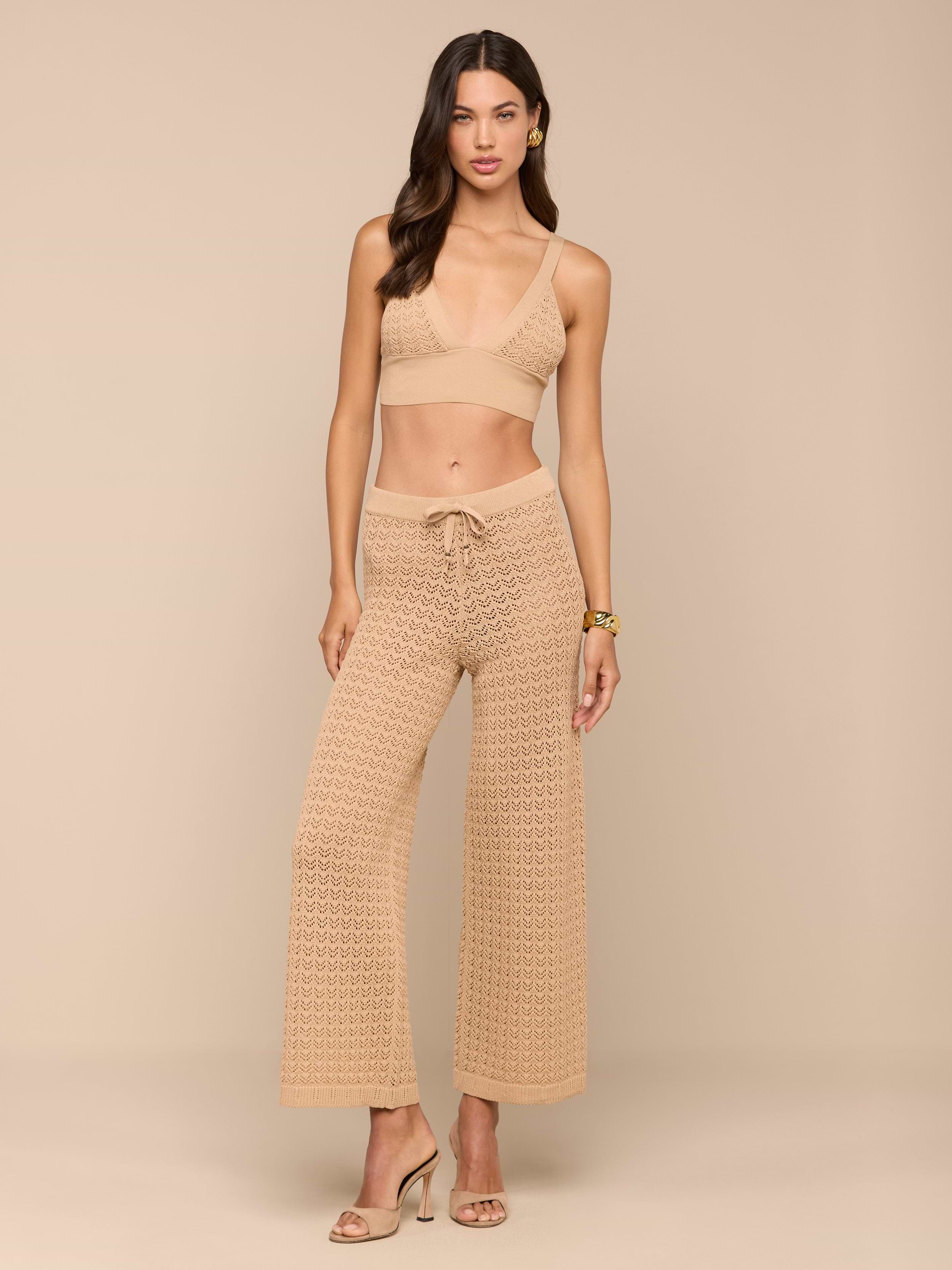 Une femme se tient debout sur un fond beige, vêtue dune bralette en crochet beige clair et dun ensemble pantalon en maille Pointelle Gaelle assortis, avec des talons ouverts. Elle a les cheveux ondulés, des boucles doreilles et un bracelet - parfait pour les vacances.