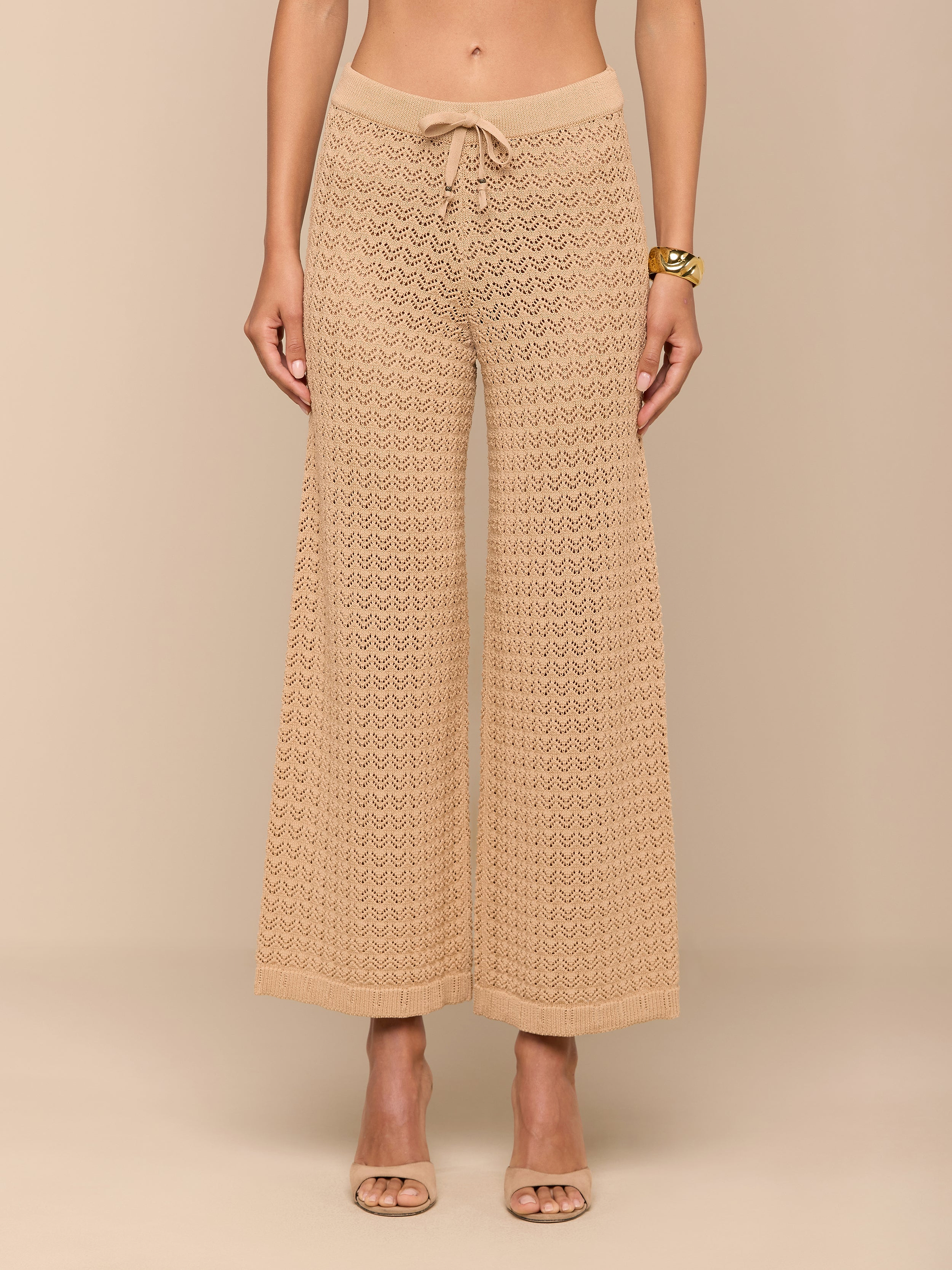 Une femme aux pieds nus présente le pantalon Gaelle Pointelle Knit Pant en beige, avec une texture en crochet, une silhouette à jambes larges et un cordon de serrage à la taille. Elle porte un bracelet en or ; le haut de son corps nest pas visible. Ce pantalon léger est prêt pour les vacances.