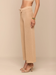 Une femme se tient debout sur un fond beige, vêtue dun pantalon Gaelle Pointelle à taille haute et à jambe large avec un lien de serrage, associé à un crop top assorti, à des mules à talons nude et à un bracelet en or.
