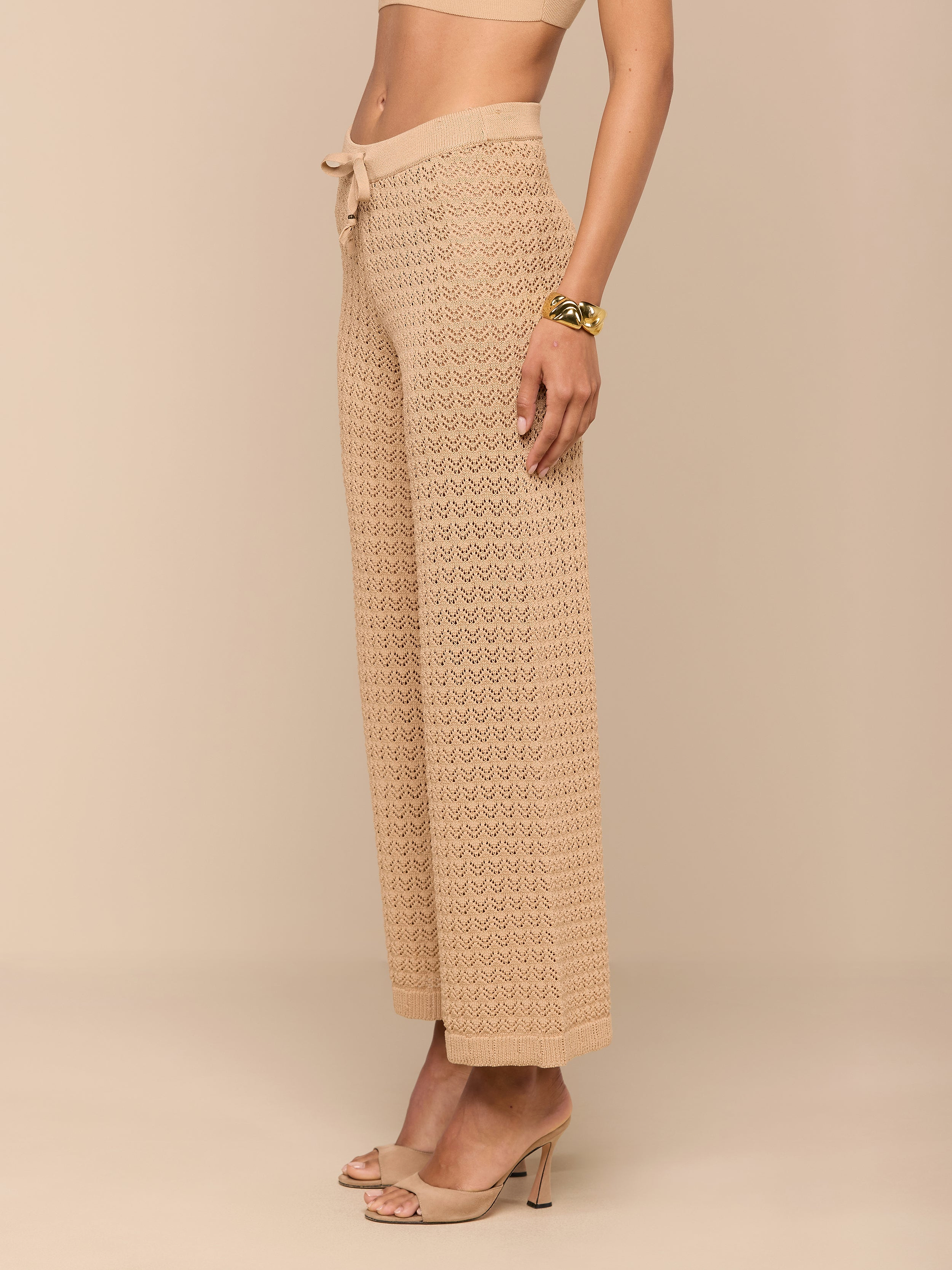 Une femme se tient debout sur un fond beige, vêtue dun pantalon Gaelle Pointelle à taille haute et à jambe large avec un lien de serrage, associé à un crop top assorti, à des mules à talons nude et à un bracelet en or.