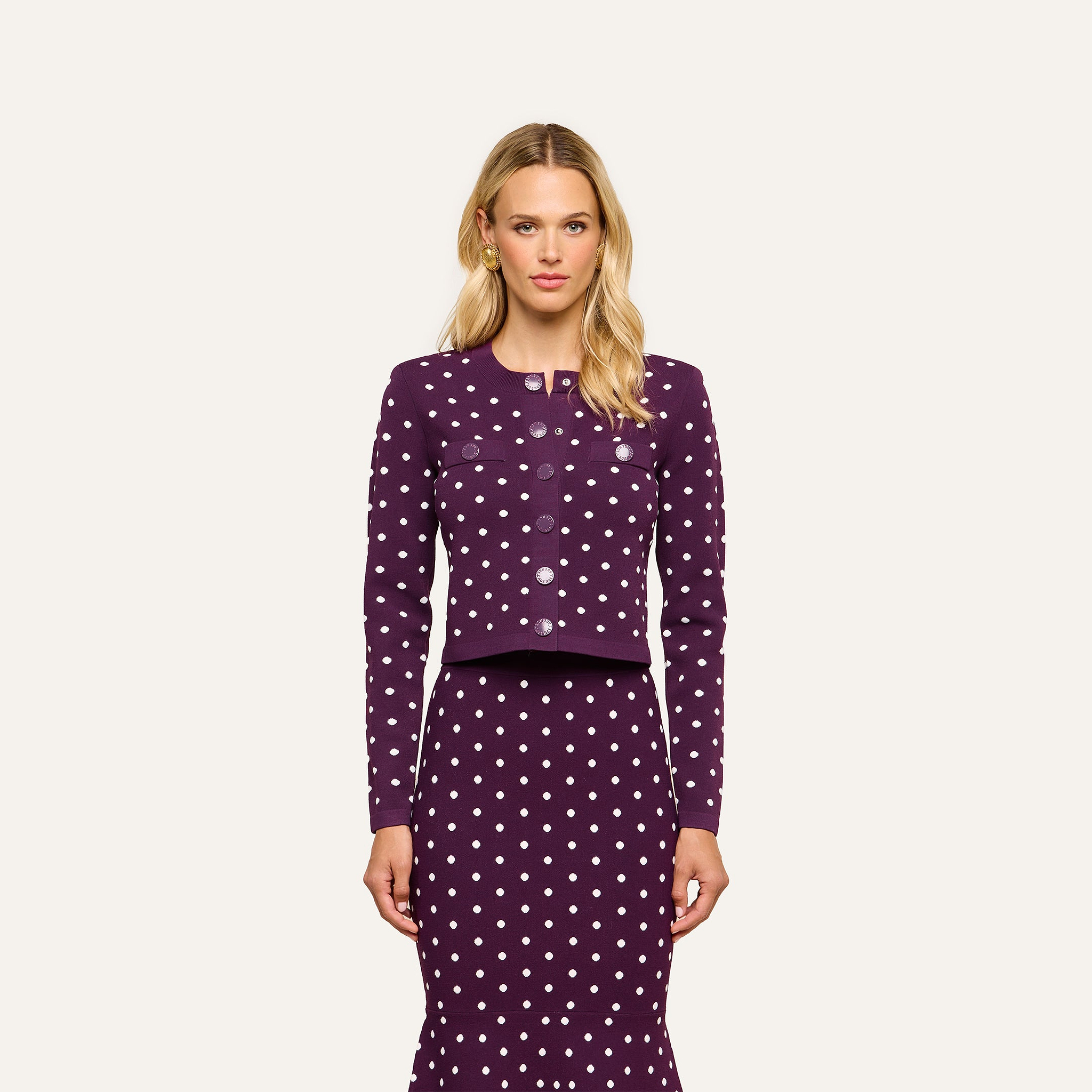 A woman wears the Kota Cardigan Top, a dark purple, long-sleeved piece with polka dots, along with a matching skirt as she stands facing forward against a plain light background.