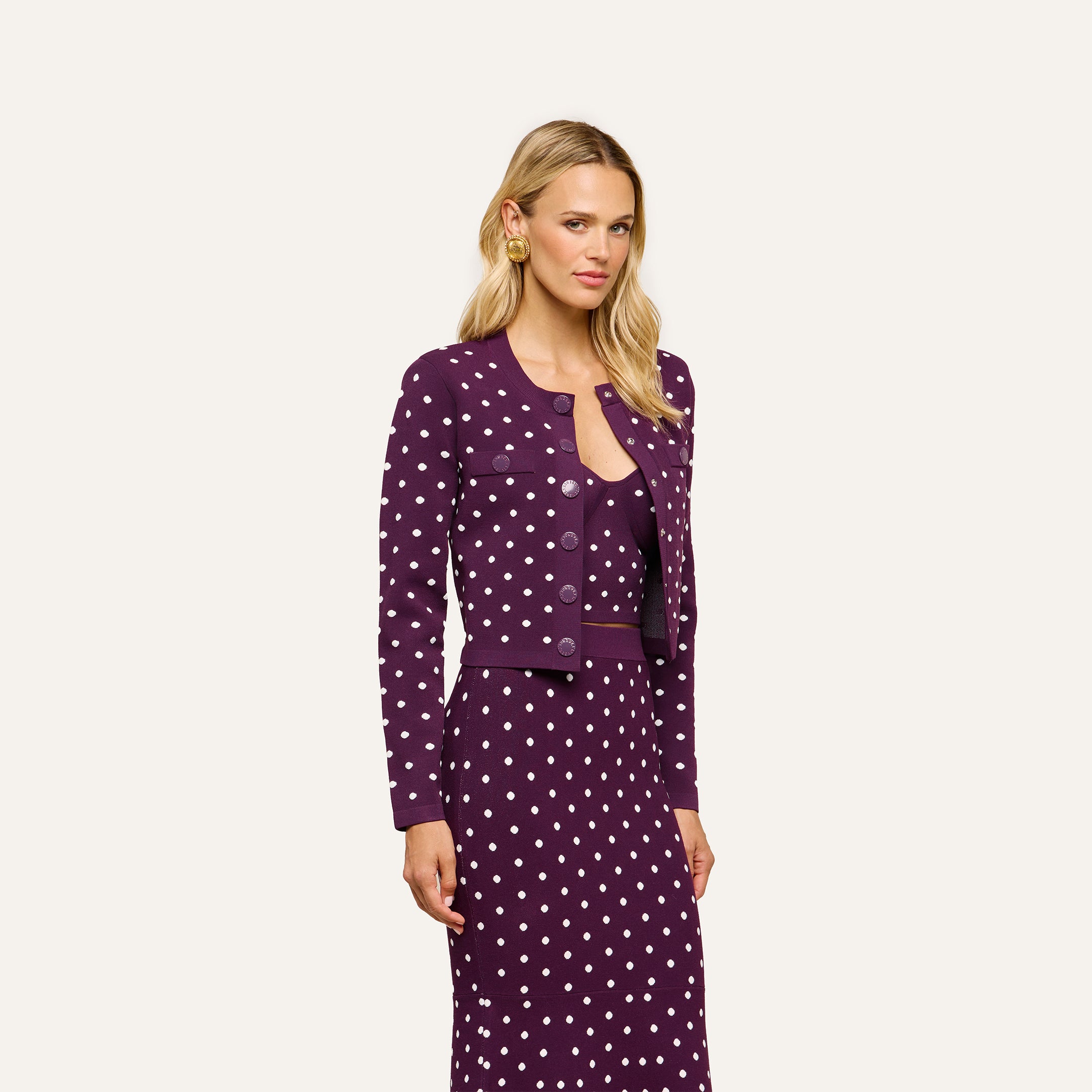 A woman stands against a plain, light background wearing the Kota Cardigan Top—a dark purple polka dot jacket—over a matching dress. She has long blonde hair and a neutral expression.
