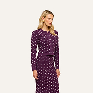A woman with long blonde hair and gold earrings stands against an off-white background, wearing the Kota Cardigan Top—a dark purple cropped cardigan and skirt set with white polka dots—while looking slightly to the side.